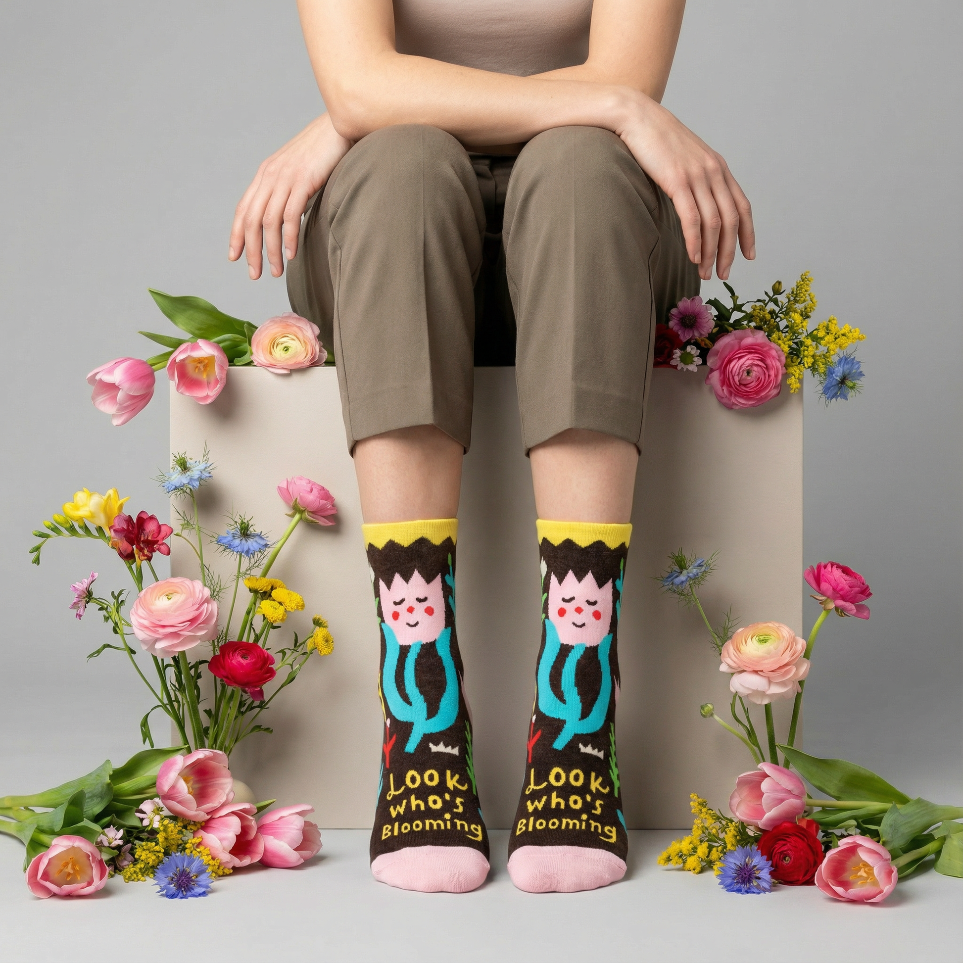 Person wearing socks with cartoon characters and text, sitting on a box with flowers around.