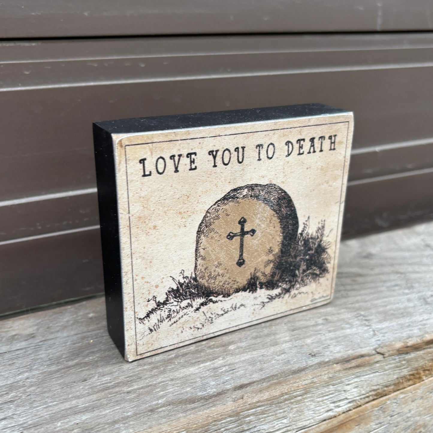 Decorative sign with 'Love You to Death' text and gravestone illustration on a wooden surface.