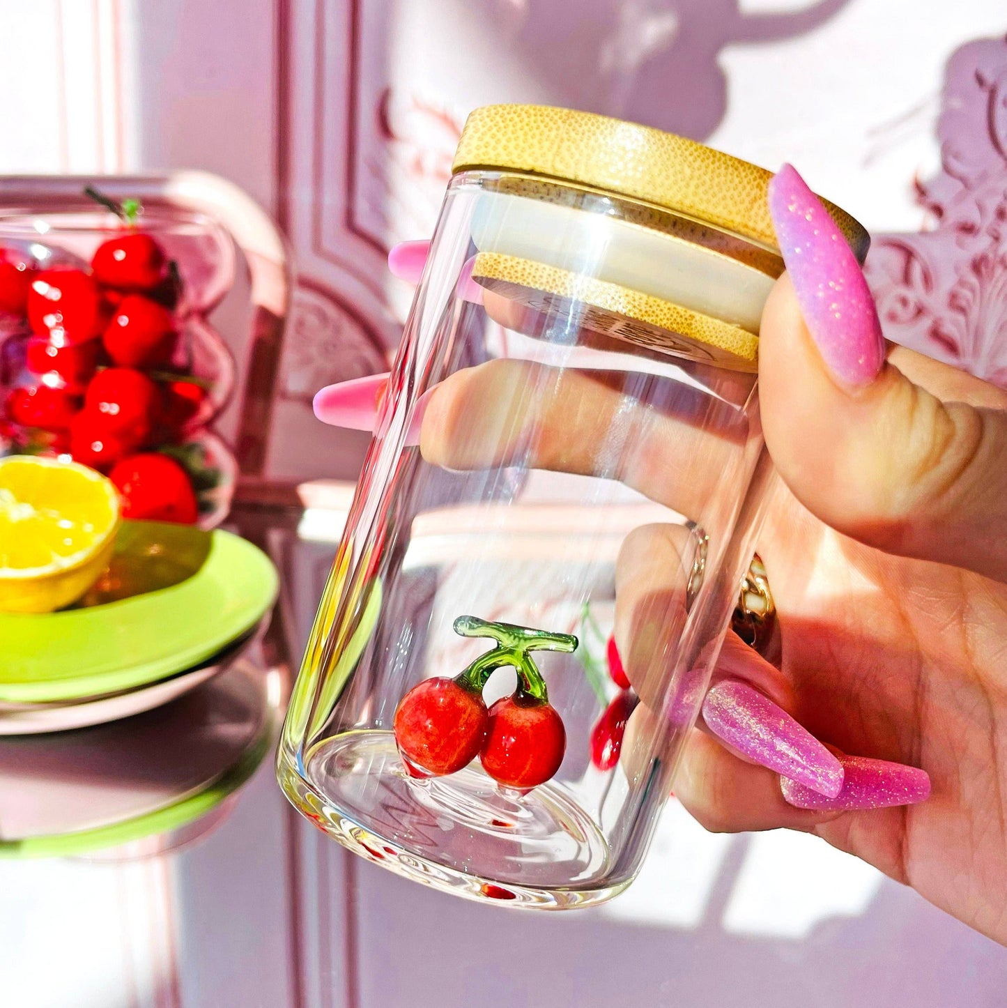 Glass jar with cherry design held by a hand with pink nail polish, blurred background