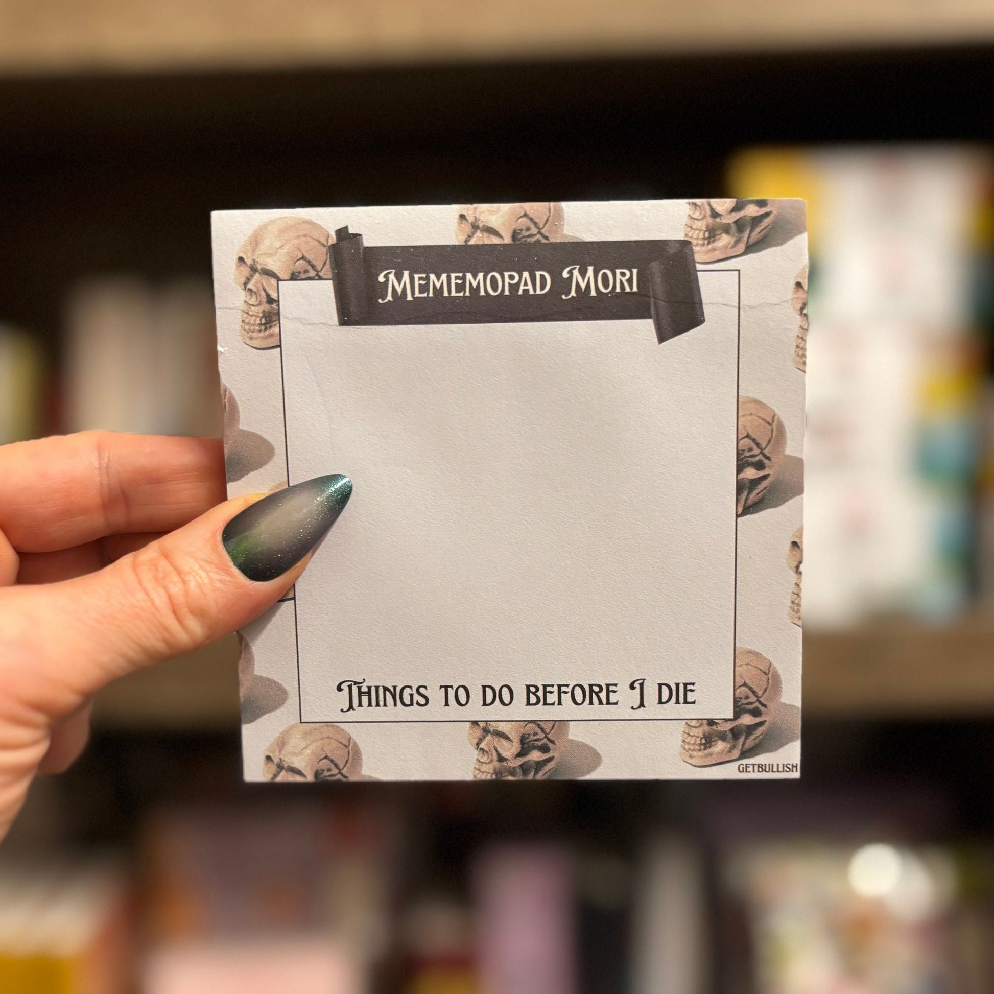 Hand holding a notepad titled 'MEMEMOPAD MORI Things to Do Before I Die' with skull designs.