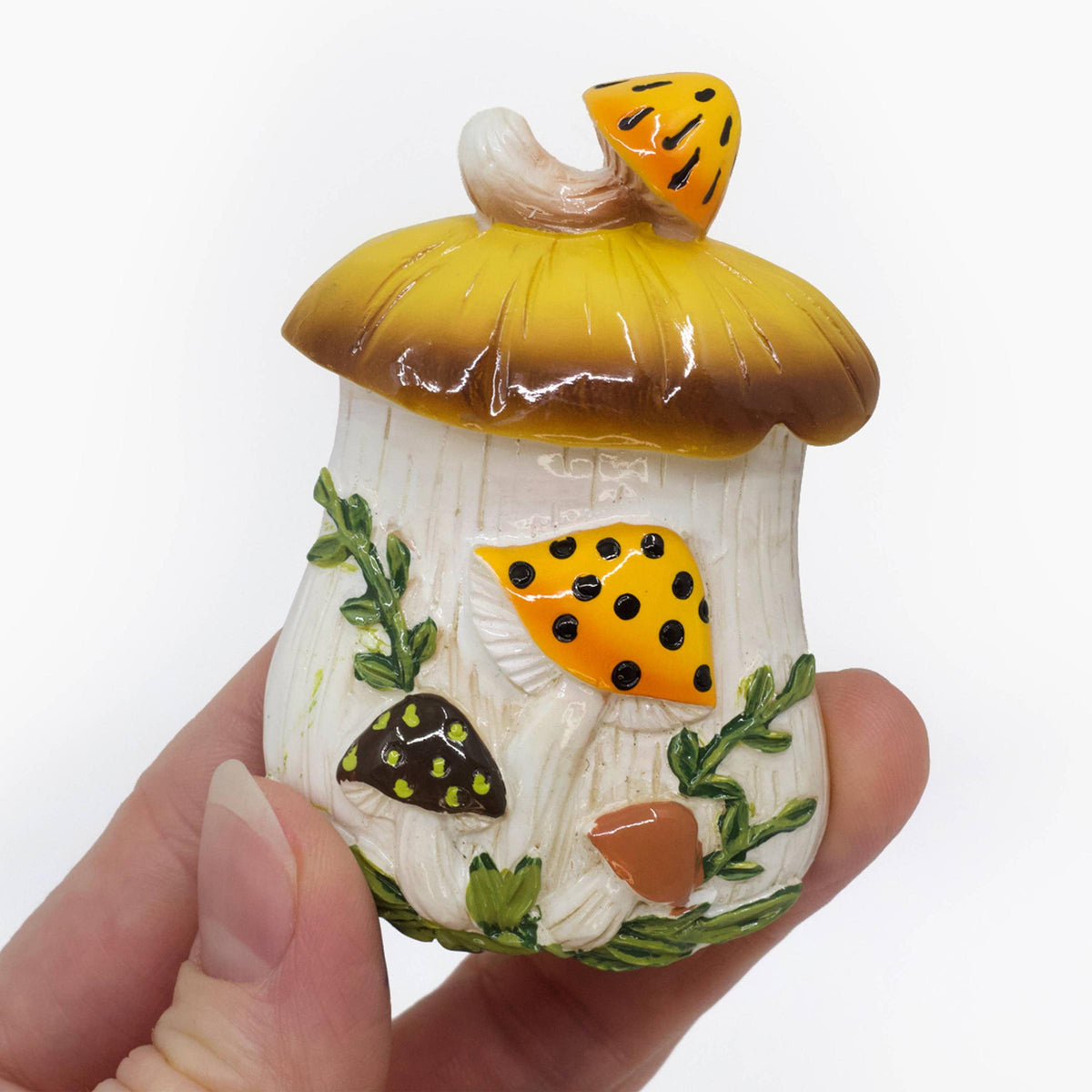 A hand holding a small decorative mushroom-shaped refrigerator magnet with a yellow and brown cap, white stem, and colorful painted details 