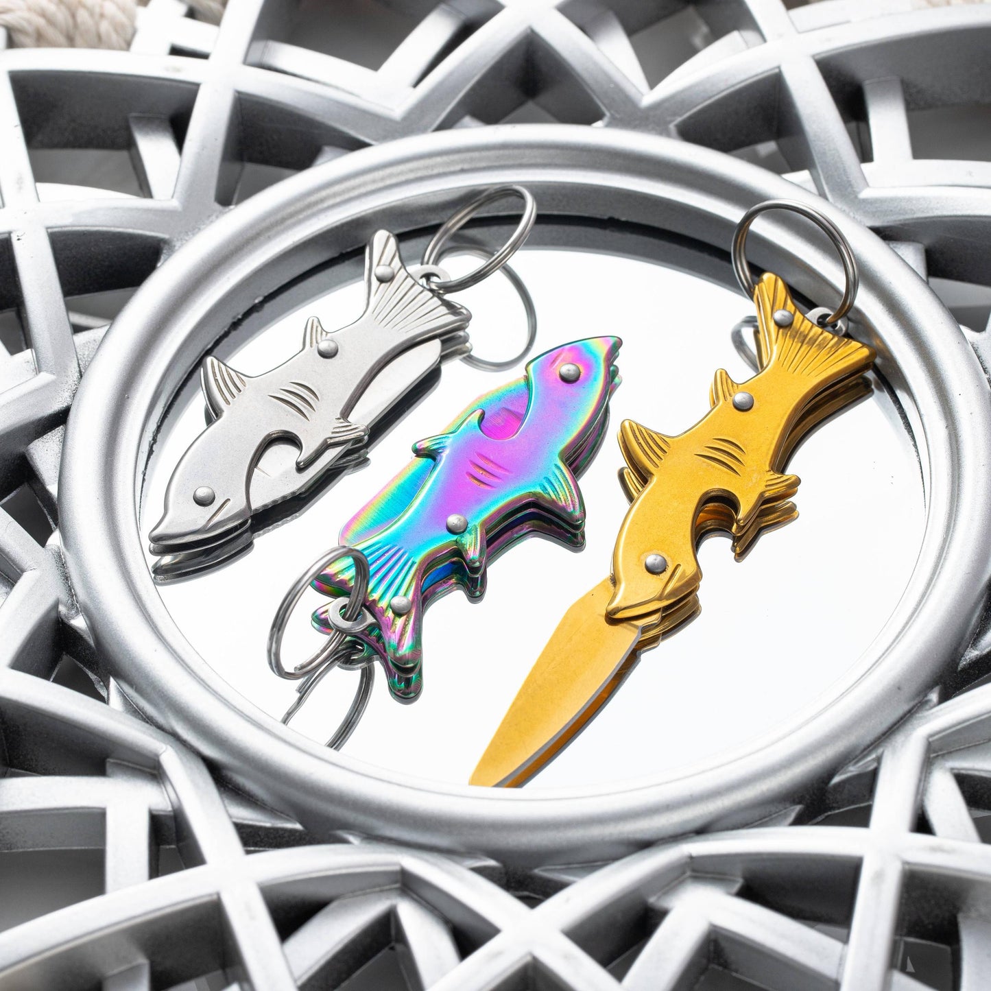 Three fish-shaped knife metal keychains in silver, iridescent rainbow, and gold finishes, displayed on a round silver tray 