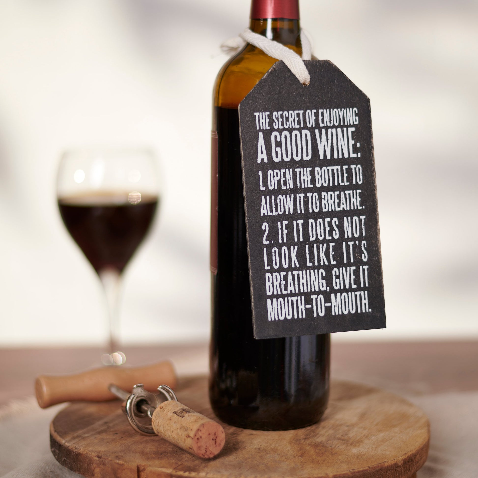 Wine bottle with a humorous tag and cork on a wooden coaster, with a glass of wine in the background.