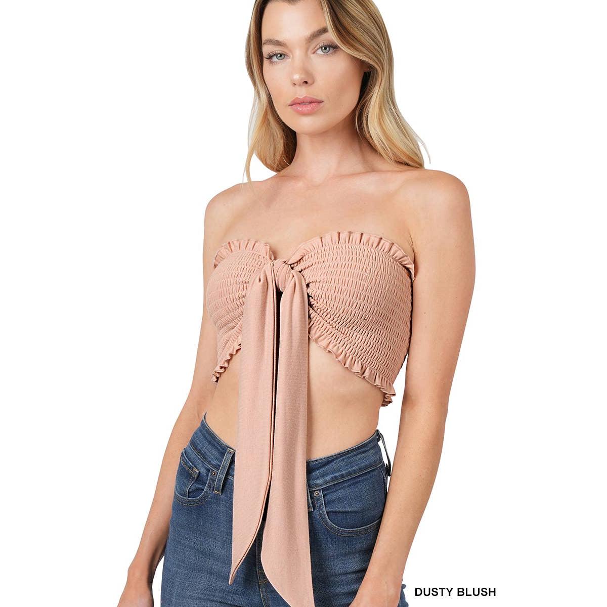 Woman wearing a dusty blush strapless top with smocked detail and tied front, paired with blue jeans on a white background.