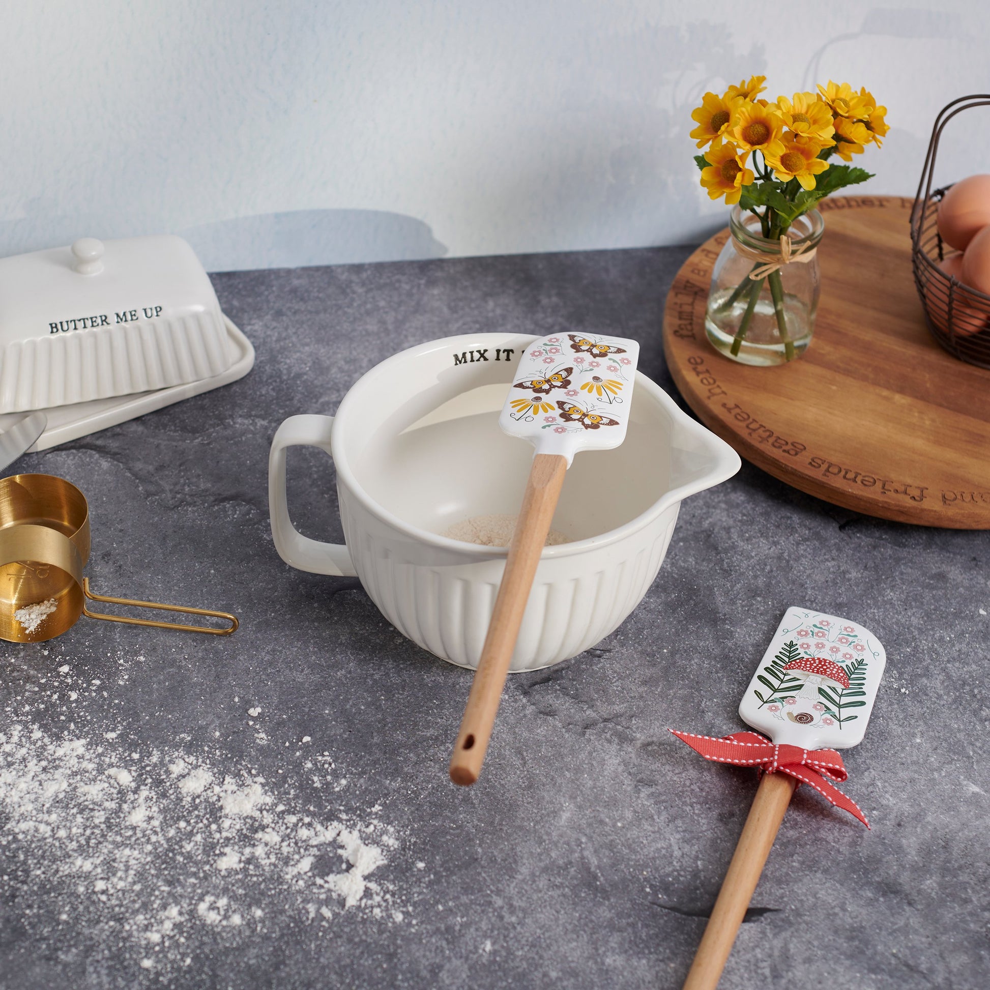 Baking utensils with decorative spatulas on a kitchen counter with flour and a vase of flowers.