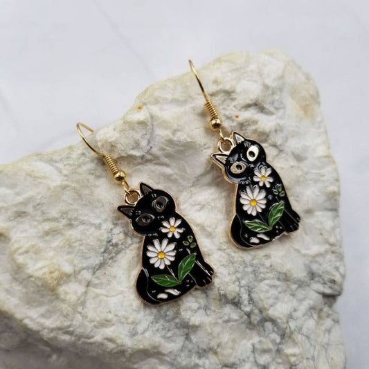 Black cat-shaped earrings with white flowers and green leaves on a stone surface