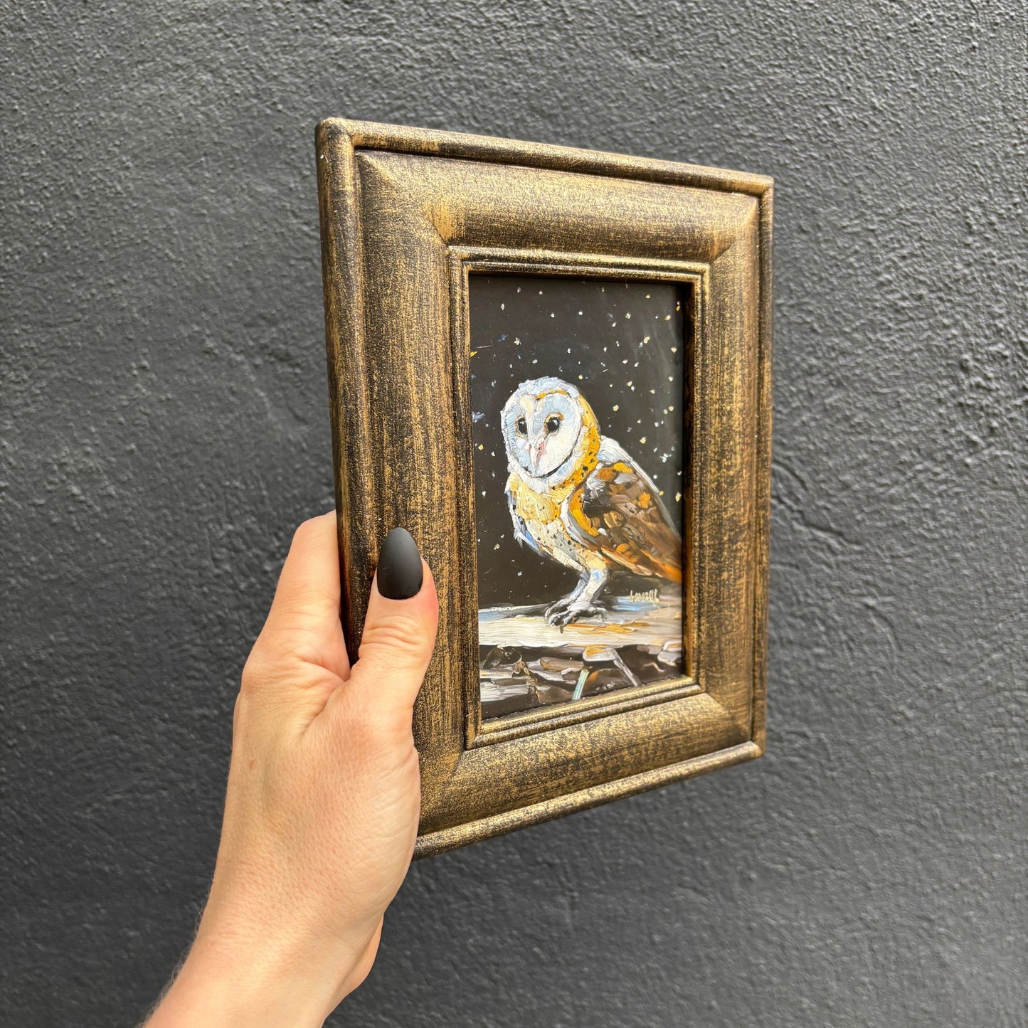 Hand holding a framed painting of an owl against a dark background
