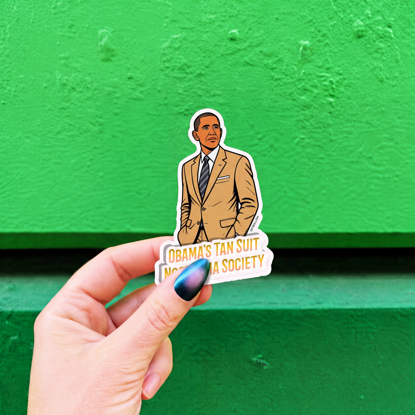 A hand with blue nail polish holds a die-cut sticker featuring an illustrated portrait of Barack Obama in a tan suit, with the text 'OBAMA'S TAN SUIT NOSTALGIA SOCIETY', against a bright green textured background.