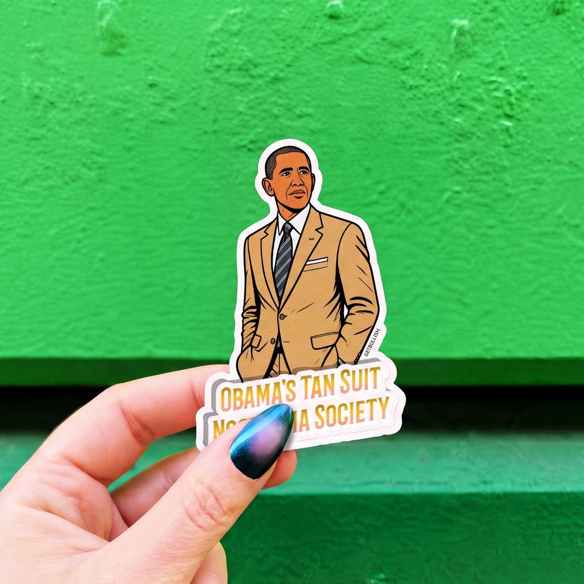 A hand with blue nail polish holds a die-cut sticker featuring an illustrated portrait of Barack Obama in a tan suit, with the text 'OBAMA'S TAN SUIT NOSTALGIA SOCIETY', against a bright green textured background.