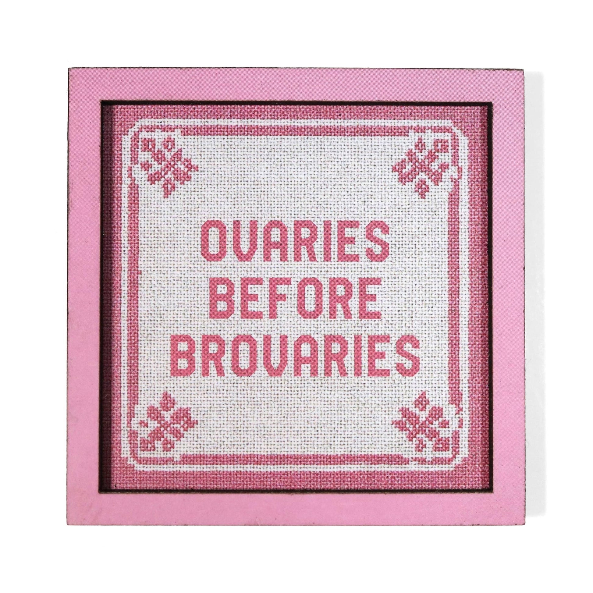 A square framed cross-stitch style magnet with pink border and text reading 'OVARIES BEFORE BROVARIES'