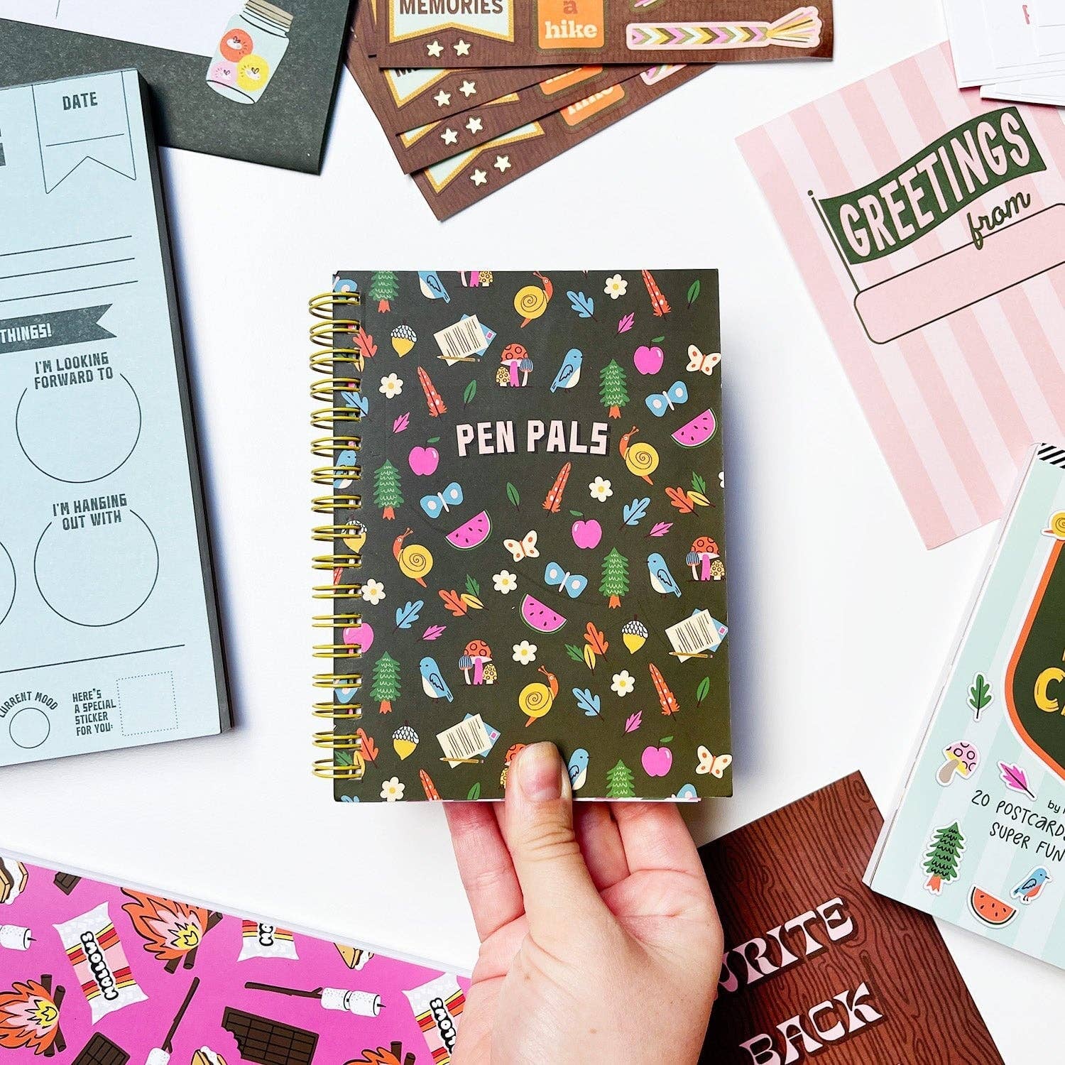 Notebook with 'Pen Pals' design held by a hand, surrounded by other notebooks and stationery items.