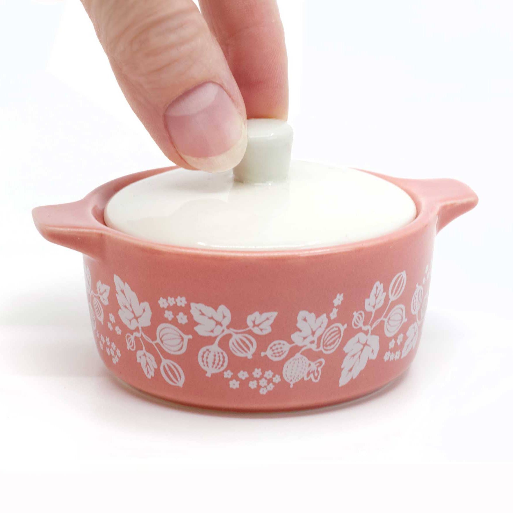 Very small pink ceramic casserole with floral patterns and a white lid being held by a hand on a white background