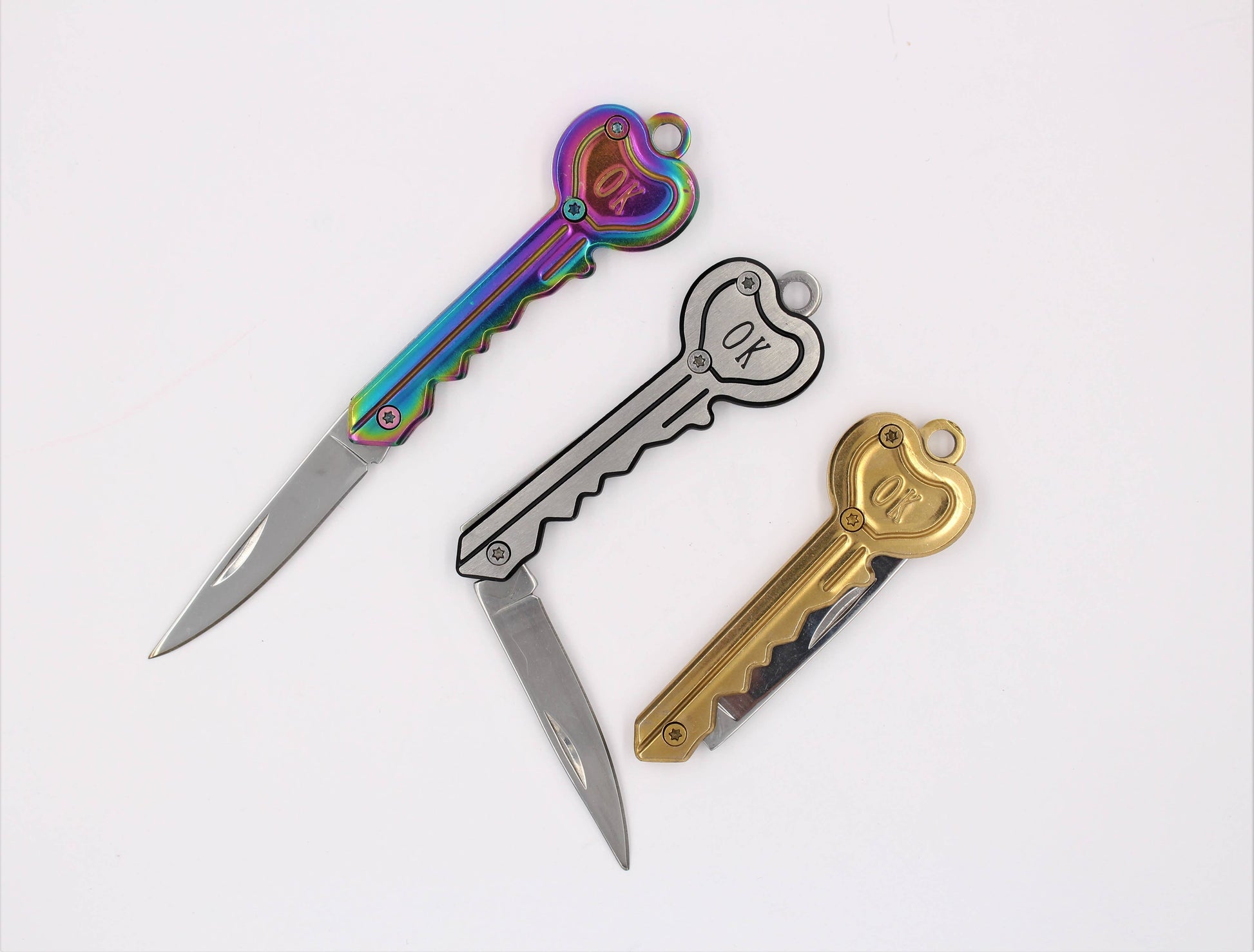 Three key-shaped folding pocket knives with heart-shaped keys. The knives feature different color options: one has an iridescent rainbow handle, one is silver with a black outline, and one is gold.