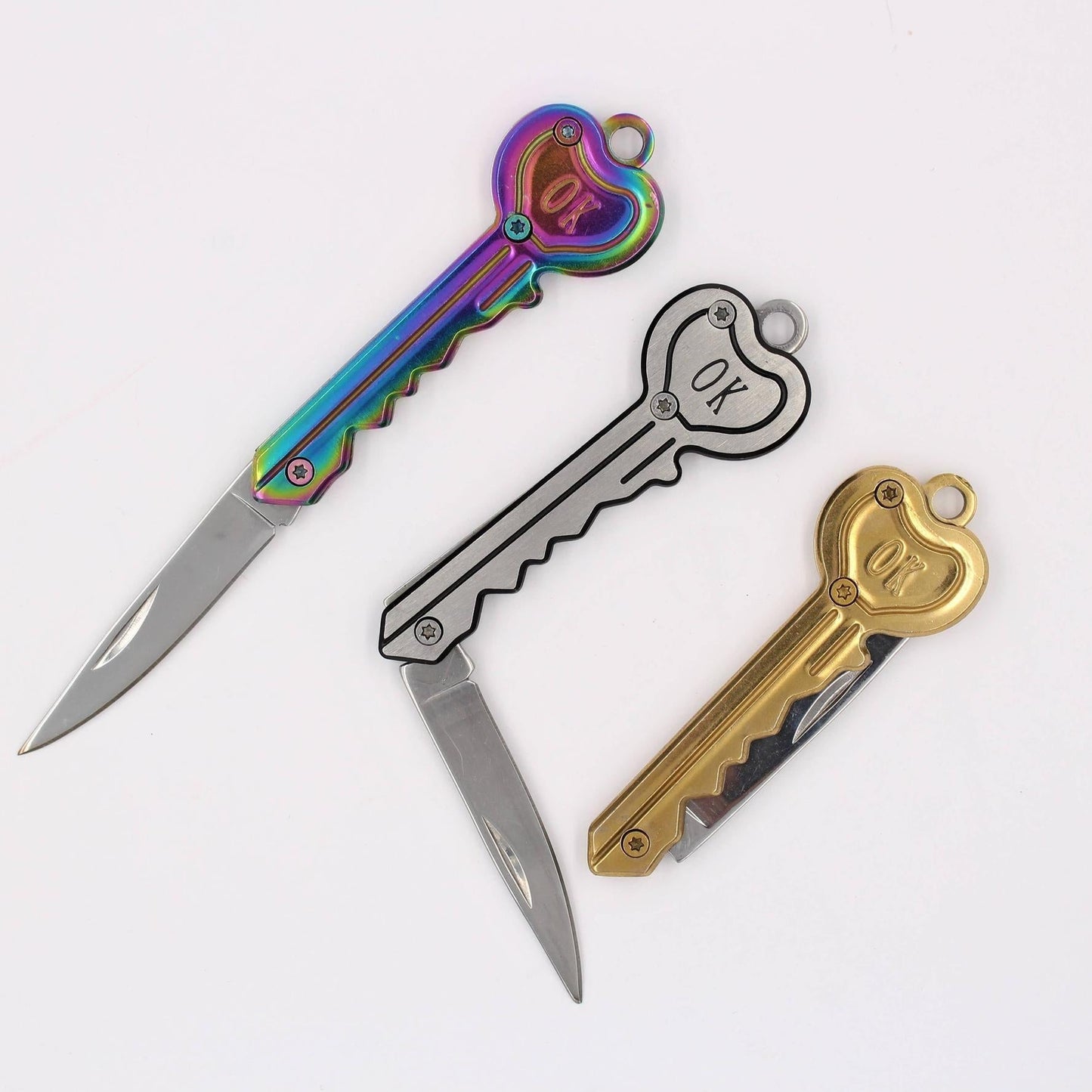 Three key-shaped folding pocket knives with heart-shaped keys. The knives feature different color options: one has an iridescent rainbow handle, one is silver with a black outline, and one is gold.