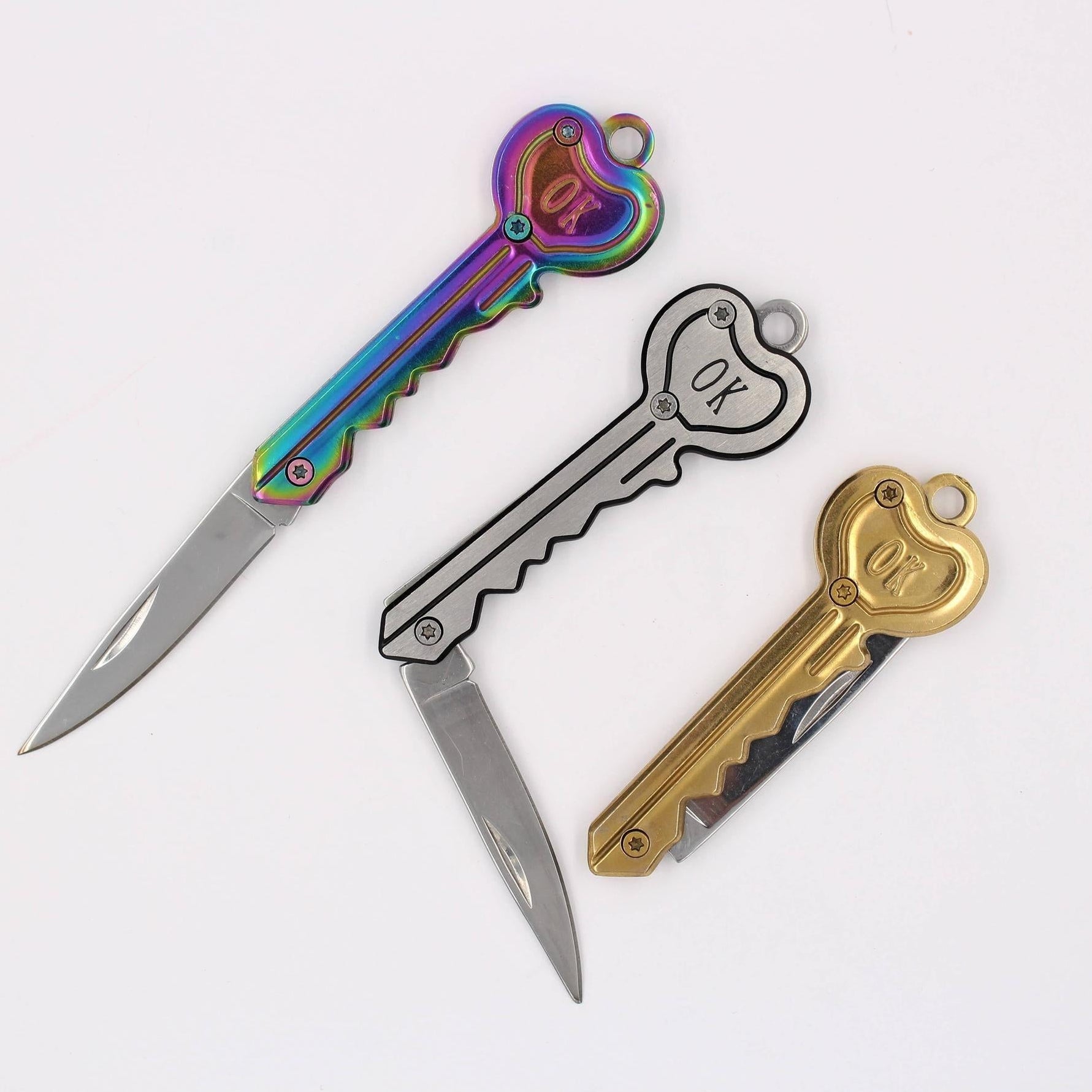 Three key-shaped folding pocket knives with heart-shaped keys. The knives feature different color options: one has an iridescent rainbow handle, one is silver with a black outline, and one is gold.