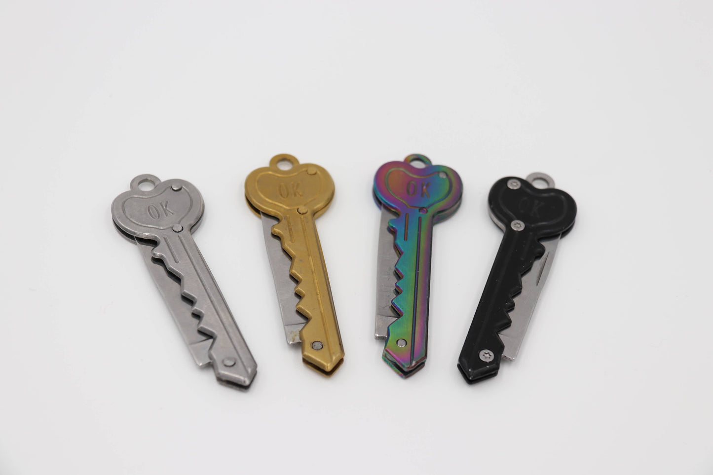 Four miniature key-shaped folding knives, showing different finish options: grey, gold, iridescent rainbow, and black.