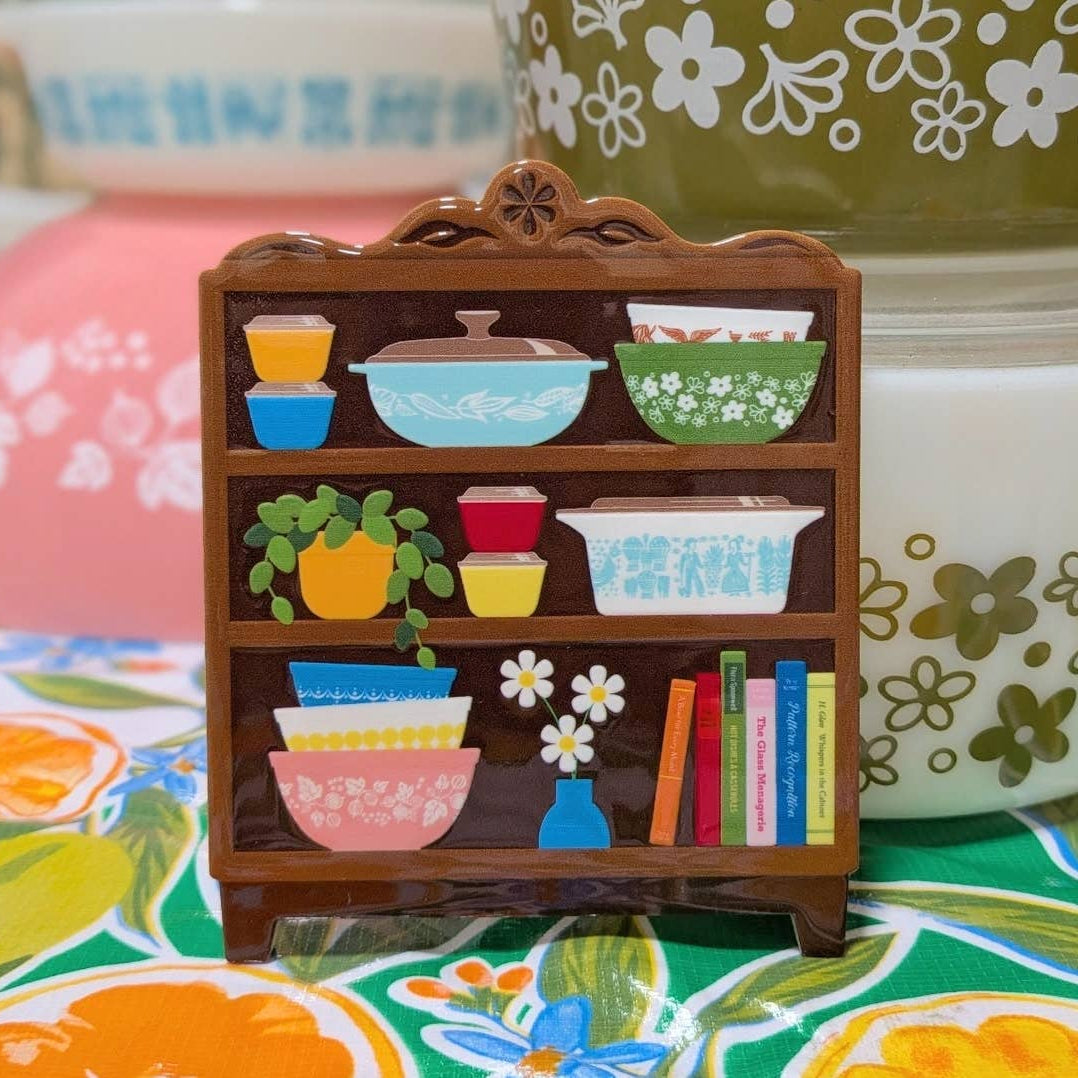 Coaster designed with miniature wooden bookshelf with Pyrex-like dishes and books on a floral-patterned surface.