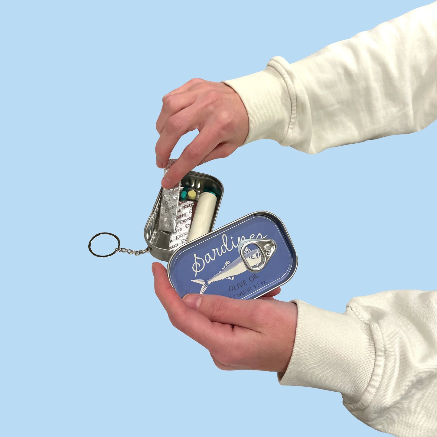 Hand opening a small metal canister-type keychain labeled 'Sardines'