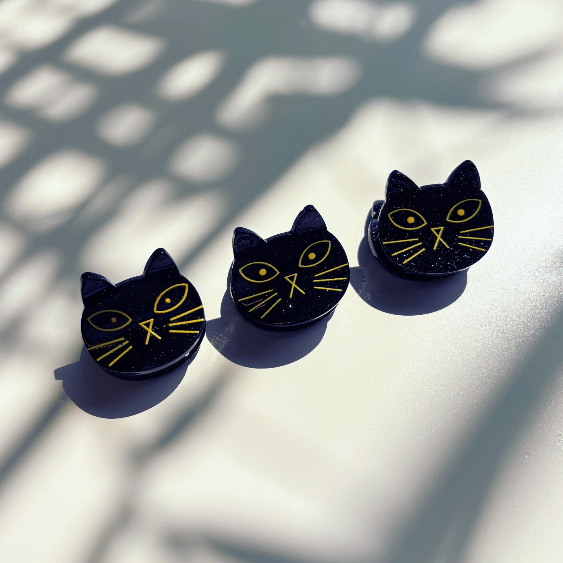 Three black cat-shaped mini hair clips with yellow details on a light surface with shadow patterns.