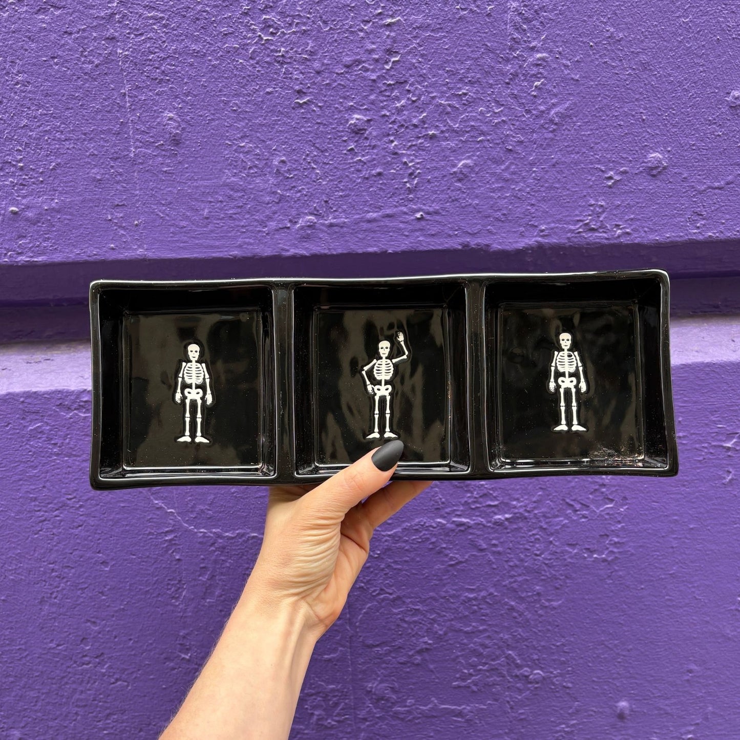 Hand holding a black tray with skeleton illustrations against a purple wall