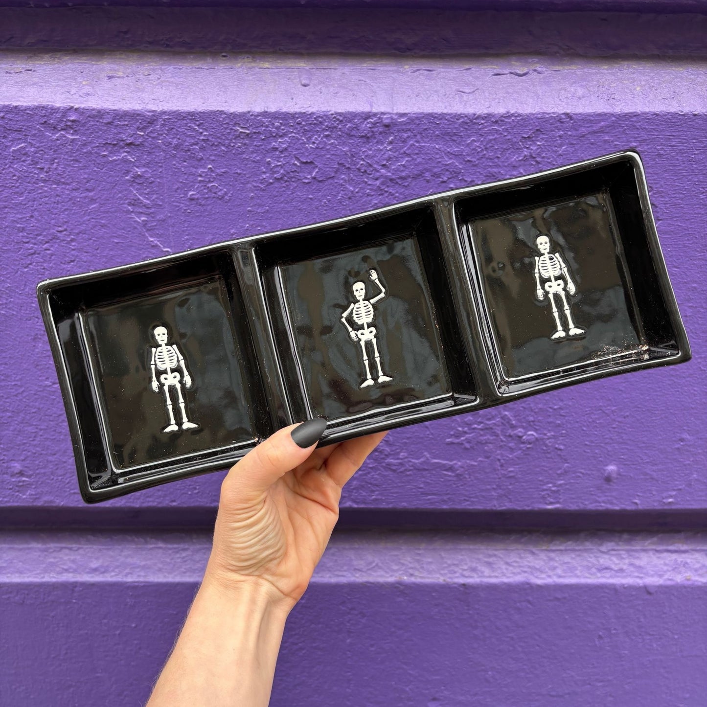 Hand holding a black tray with three small black boxes containing white skeletons against a purple wall.