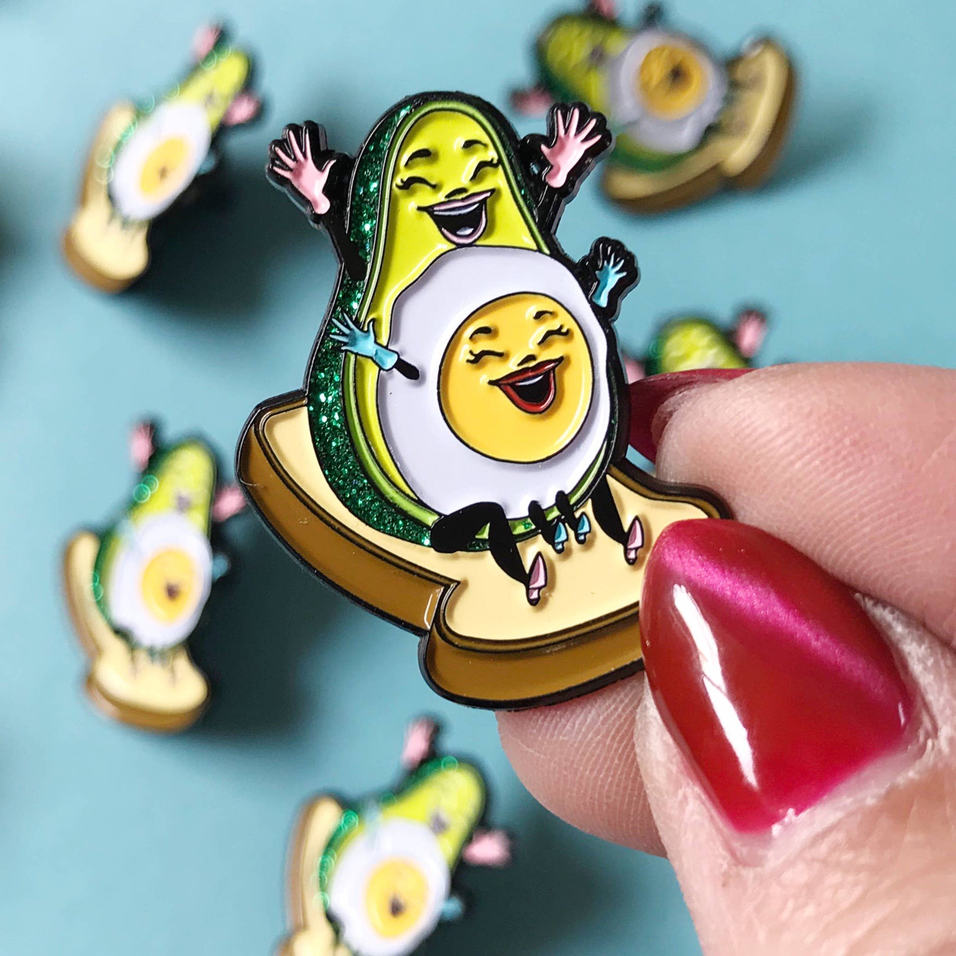 Avocado and egg character pin held by a hand with red nail polish on a blue background
