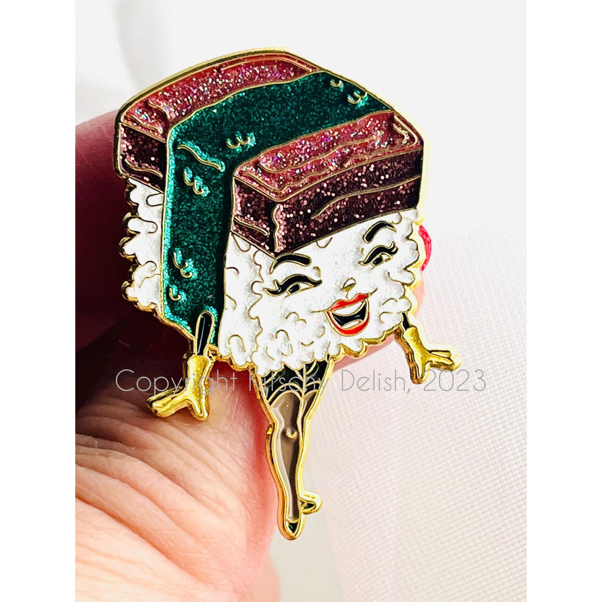 A close-up of a hand holding a colorful enamel pin shaped like a smiling sushi character with glittery green and red toppings, white rice, and gold outlines, against a light background.