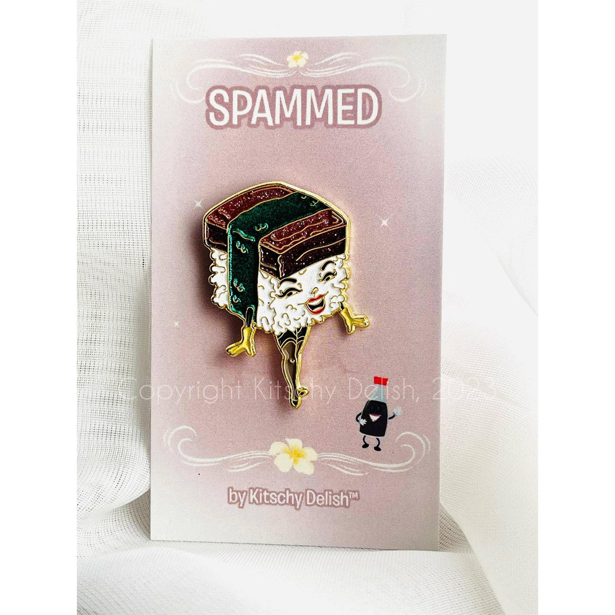 A cute spammed sushi enamel pin displayed on its pink backing card, which reads 'SPAMMED' and is branded 'by Kitschy Delish'. The pin features a smiling sushi character with white rice, green seaweed, and gold metal outlines.