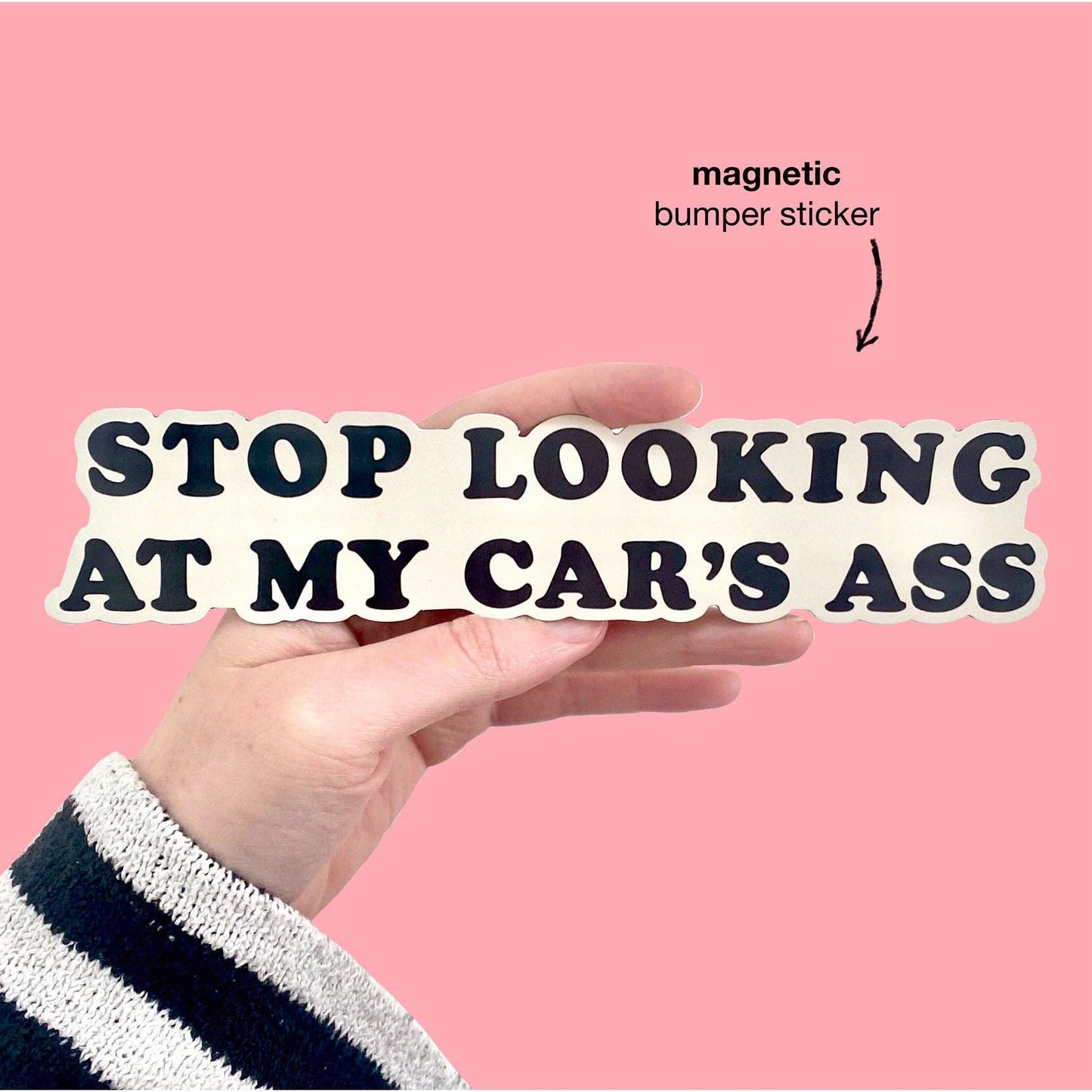 Hand holding a bumper sticker with text 'Stop looking at my car's ass' against a pink background