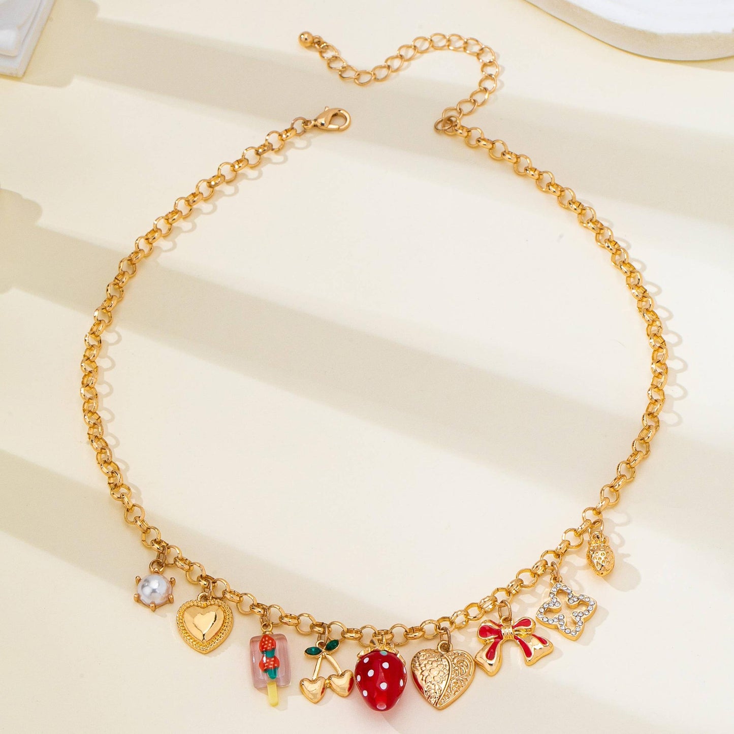 A gold-tone charm necklace with a delicate link chain and multiple small charms, including hearts, a strawberry, cherries, a bow, and a pearl-like bead