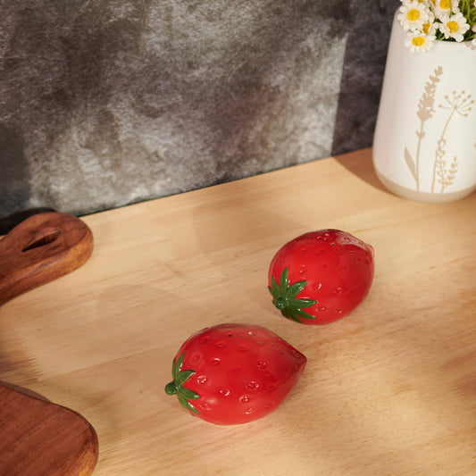 Two red strawberry-shaped salt and pepper shakers on a wooden surface with a vase in the background.