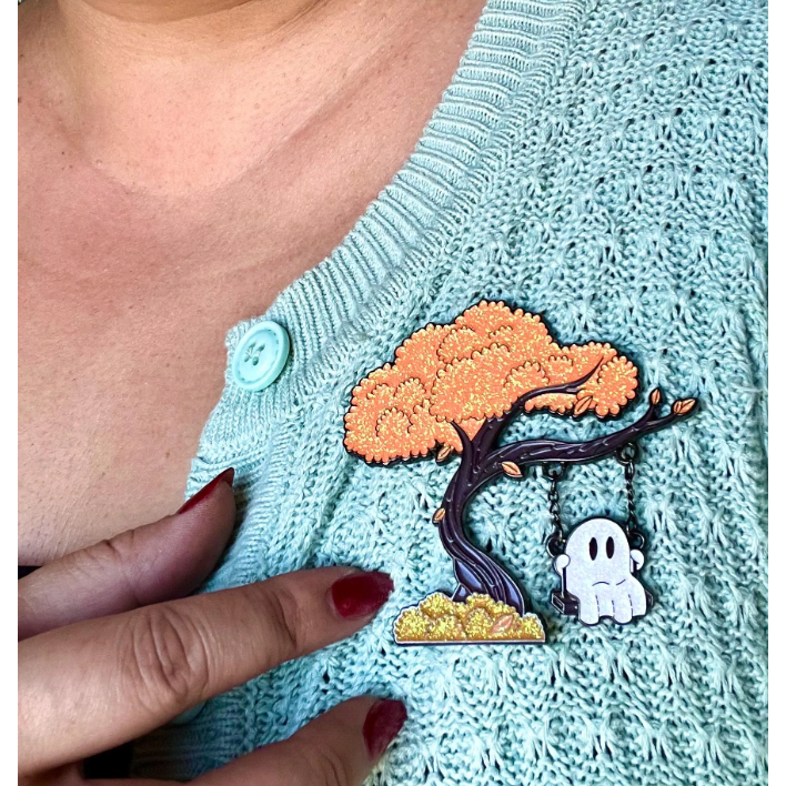 Person wearing a light blue knitted sweater with a tree and ghost enamel pin