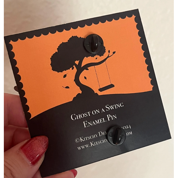 Enamel pin with 'Ghost on a Swing' design on an orange and black card held by a hand.