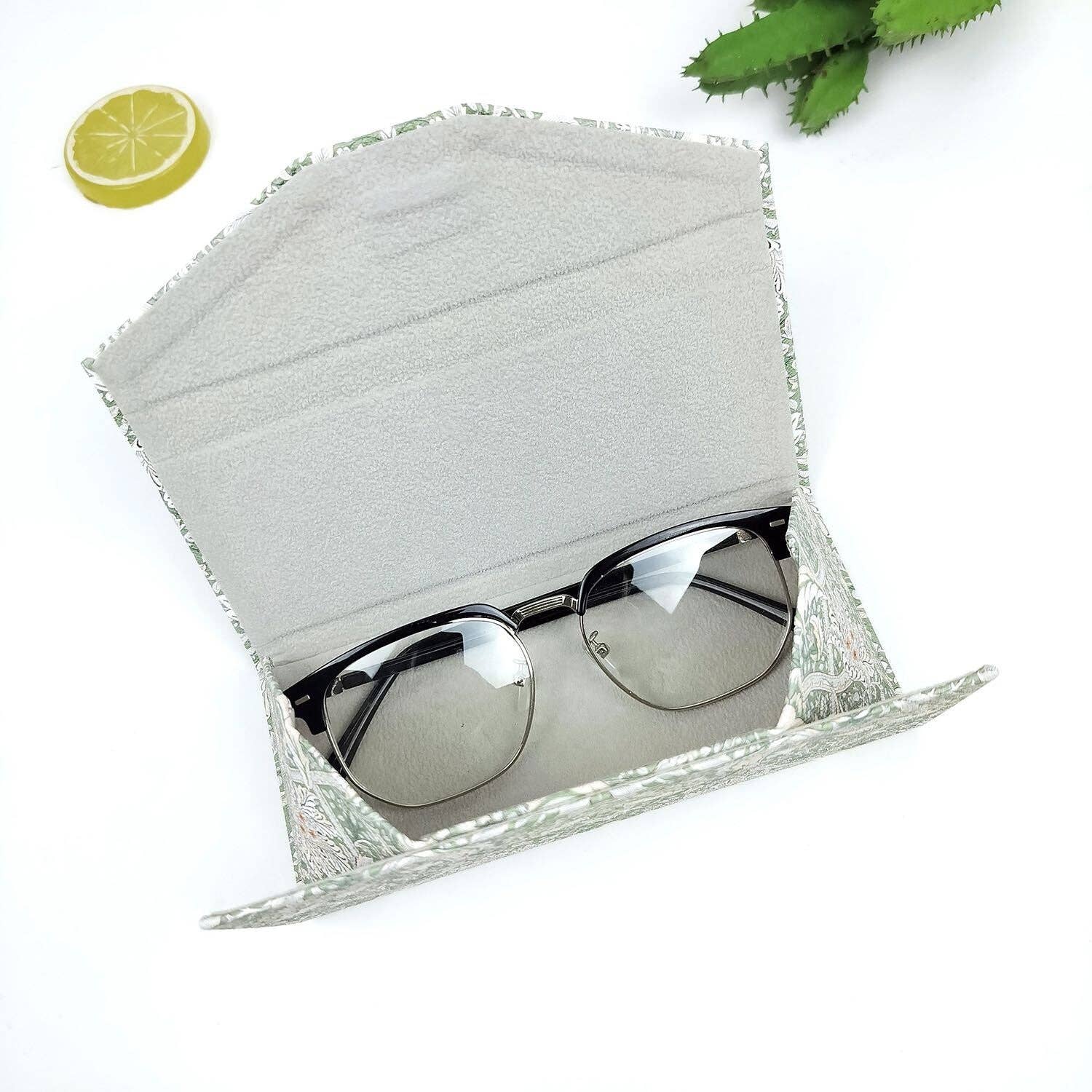 Silver sunglasses in a textured gray case on a white background with green leaves.