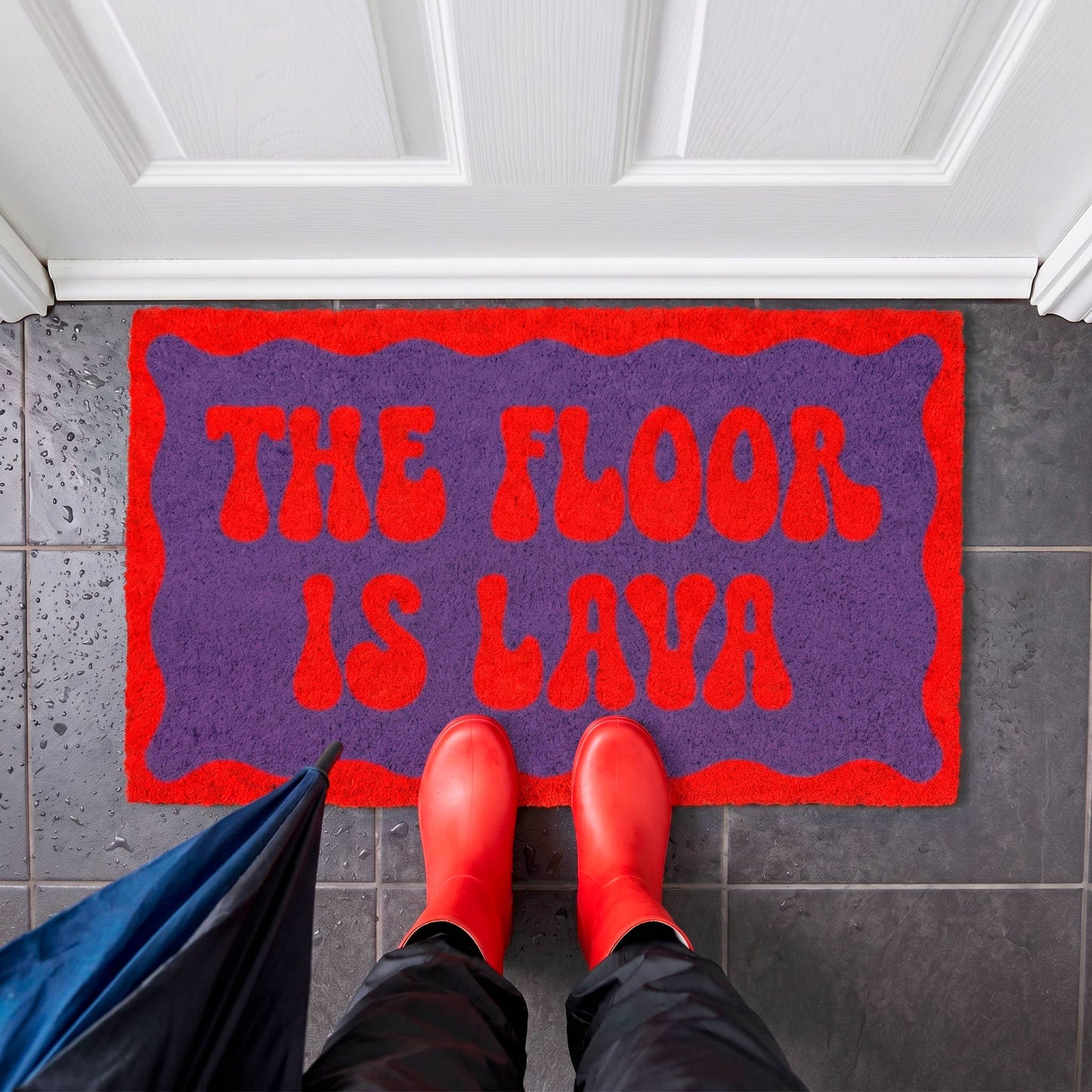 A purple and red doormat with the text 'THE FLOOR IS LAVA' in red, shown on a dark tiled entryway floor with a person's feet in red rain boots standing on it. A blue umbrella is partially visible at the lower left.