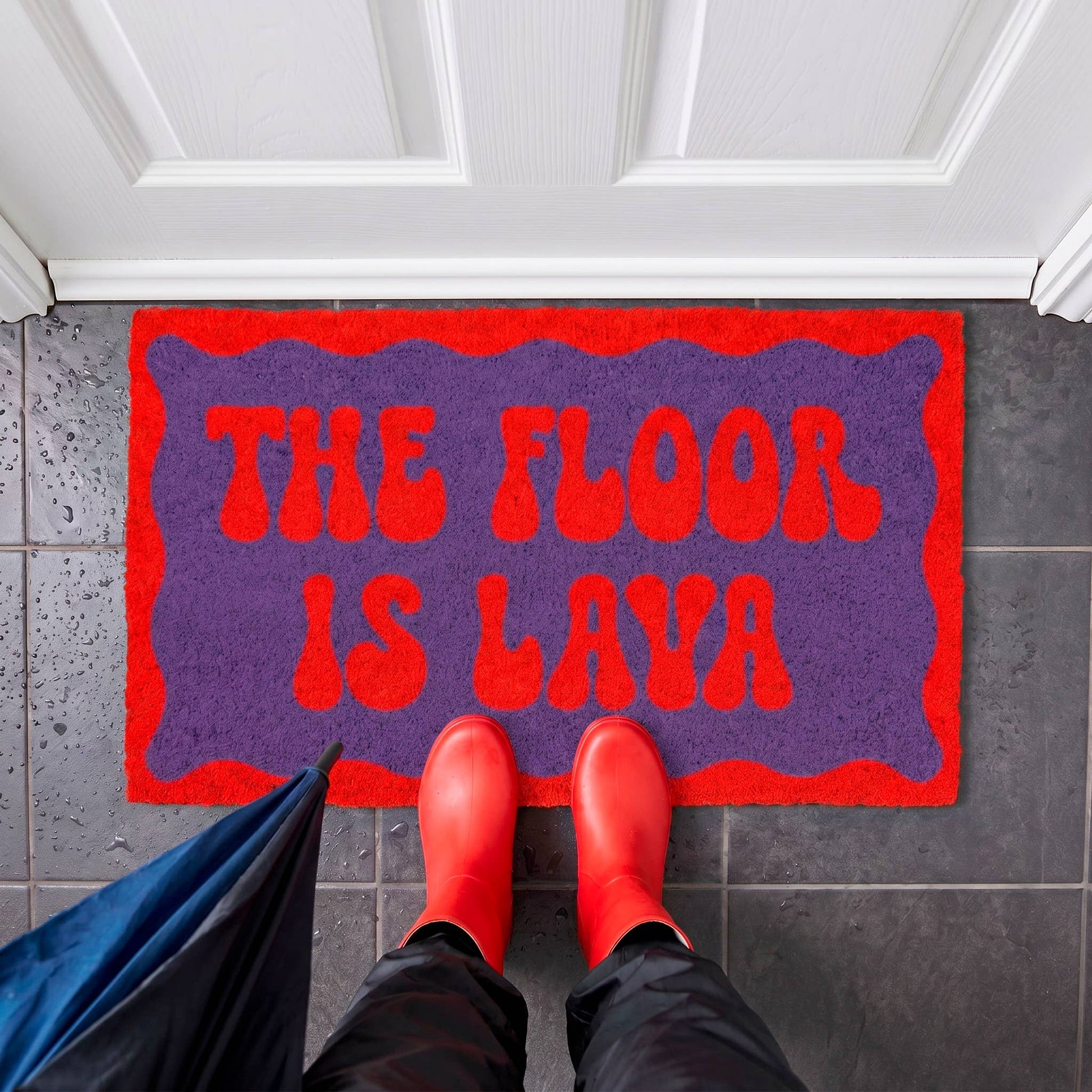 A purple and red doormat with the text 'THE FLOOR IS LAVA' in red, shown on a dark tiled entryway floor with a person's feet in red rain boots standing on it. A blue umbrella is partially visible at the lower left.
