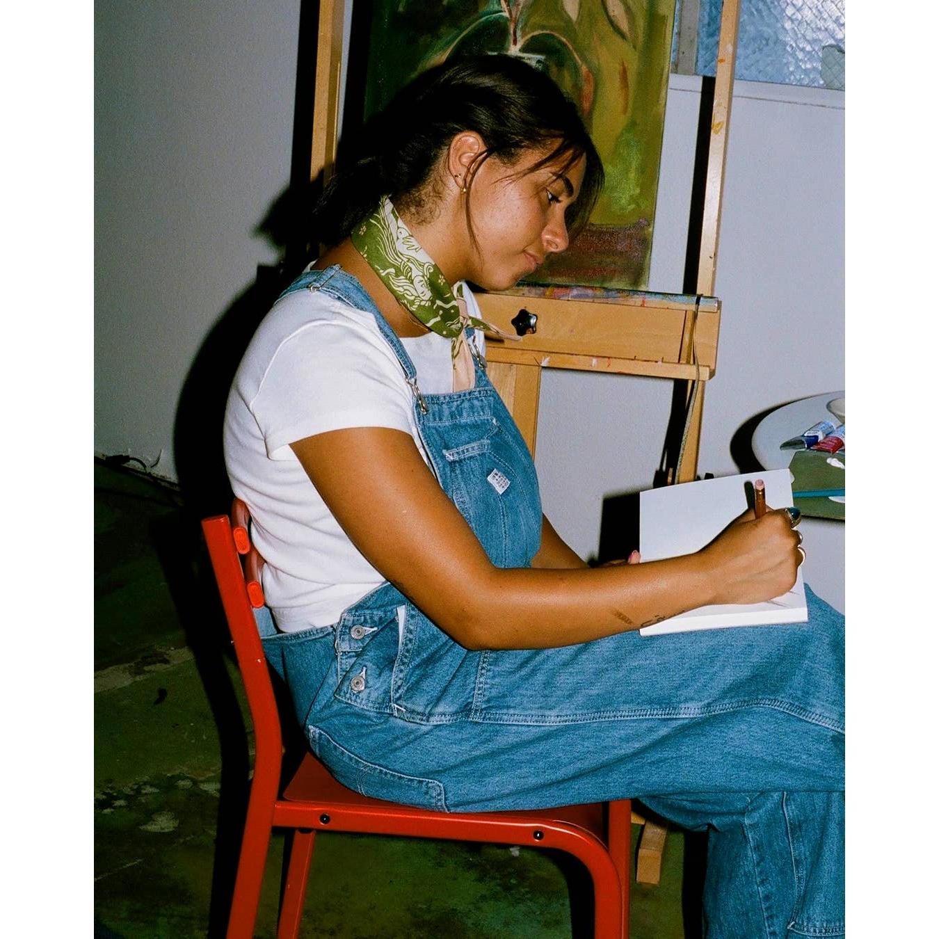 Person wearing green bandana as necklace sitting on a red chair drawing at an easel with a painting in the background.