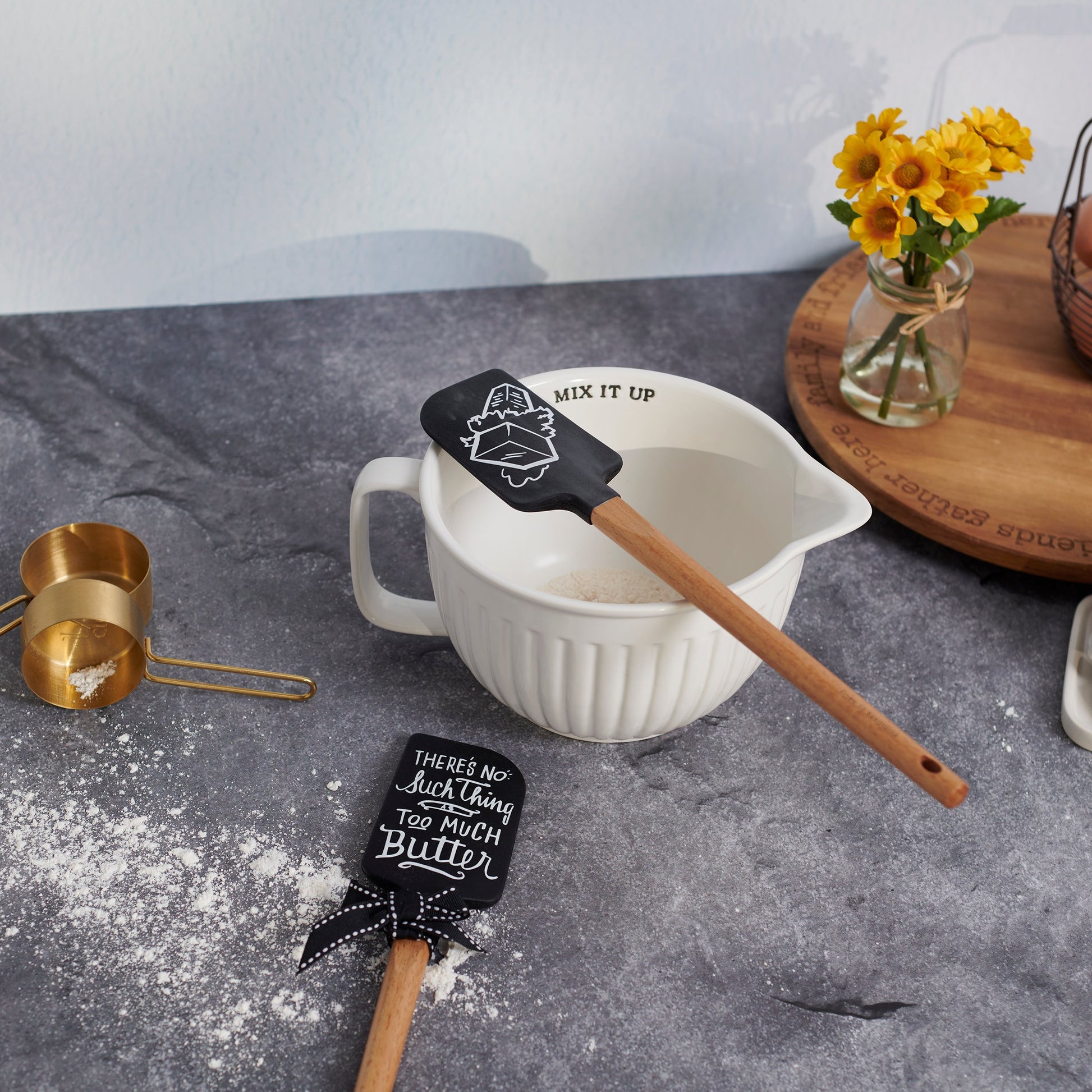 White mixing bowl with black spatulas on a gray surface with yellow flowers in the background