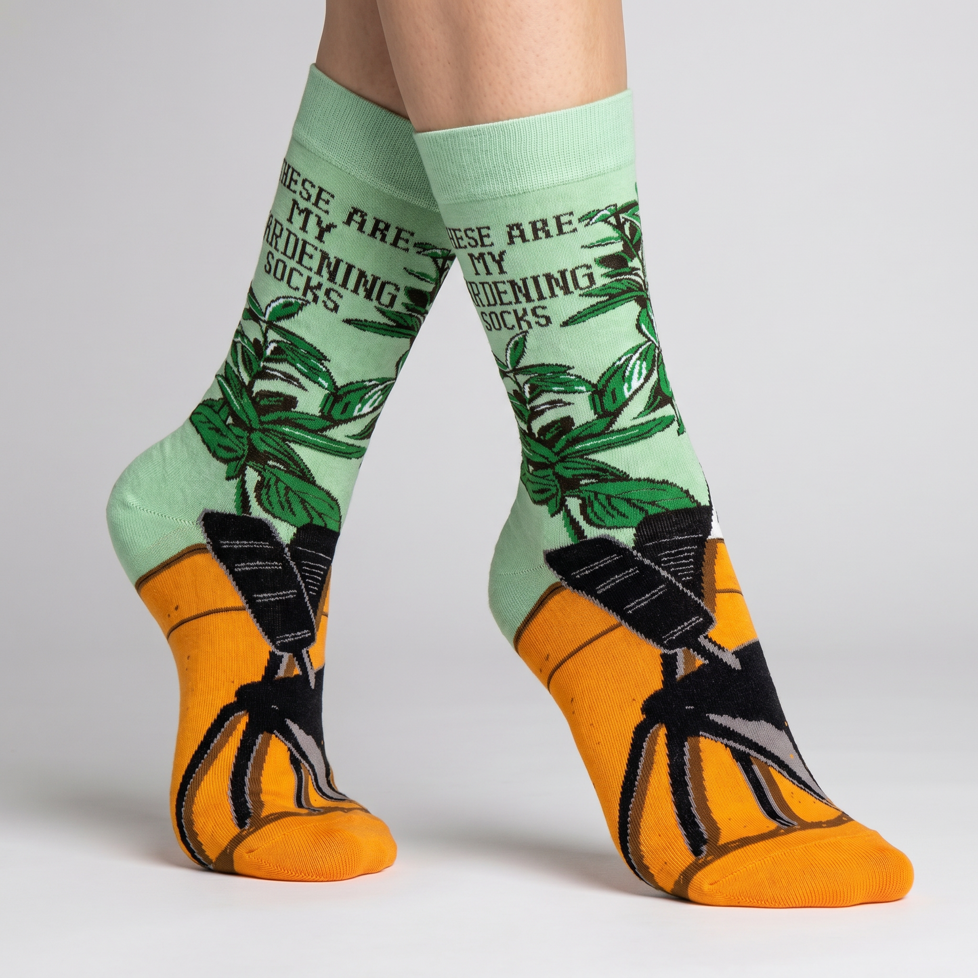 Green gardening-themed socks with text and black and orange socks worn by a person on a light gray background.
