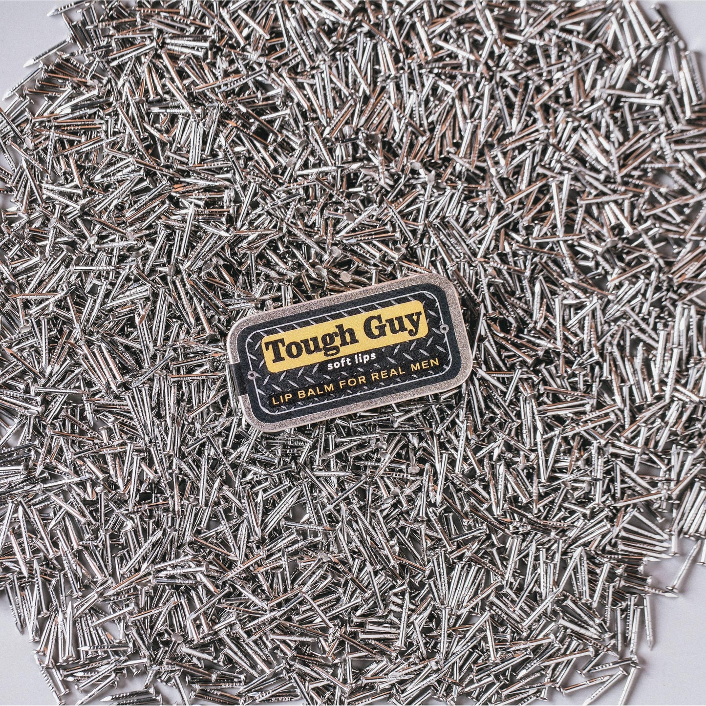 Tough Guy lip balm surrounded by small metal objects on a white background