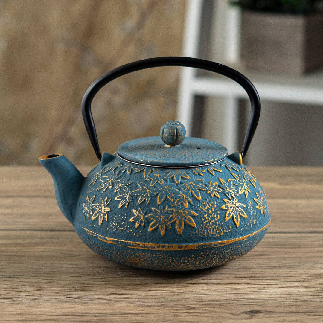 A round teal cast iron tea kettle with gold floral engravings and a black handle