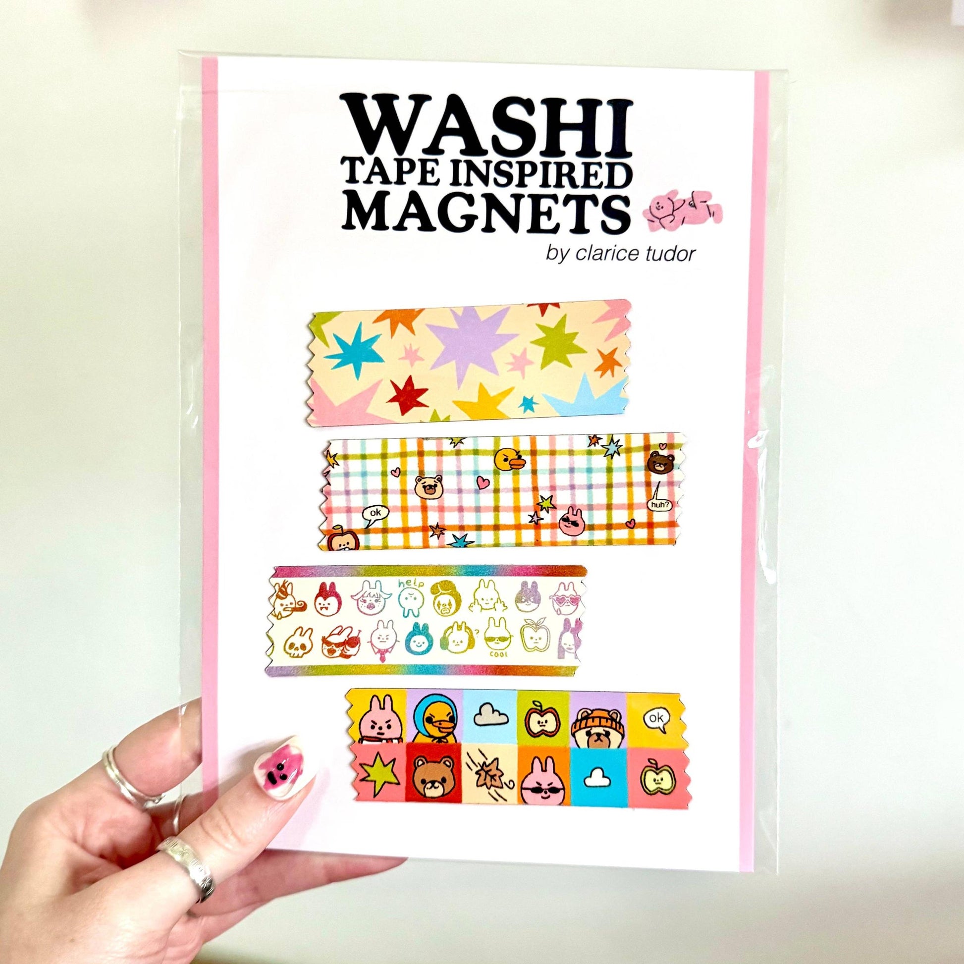 A hand holds a clear plastic sleeve containing a set of washi tape inspired magnets. The magnets feature various colorful washi tape patterns, including stars, plaid, and cartoon characters