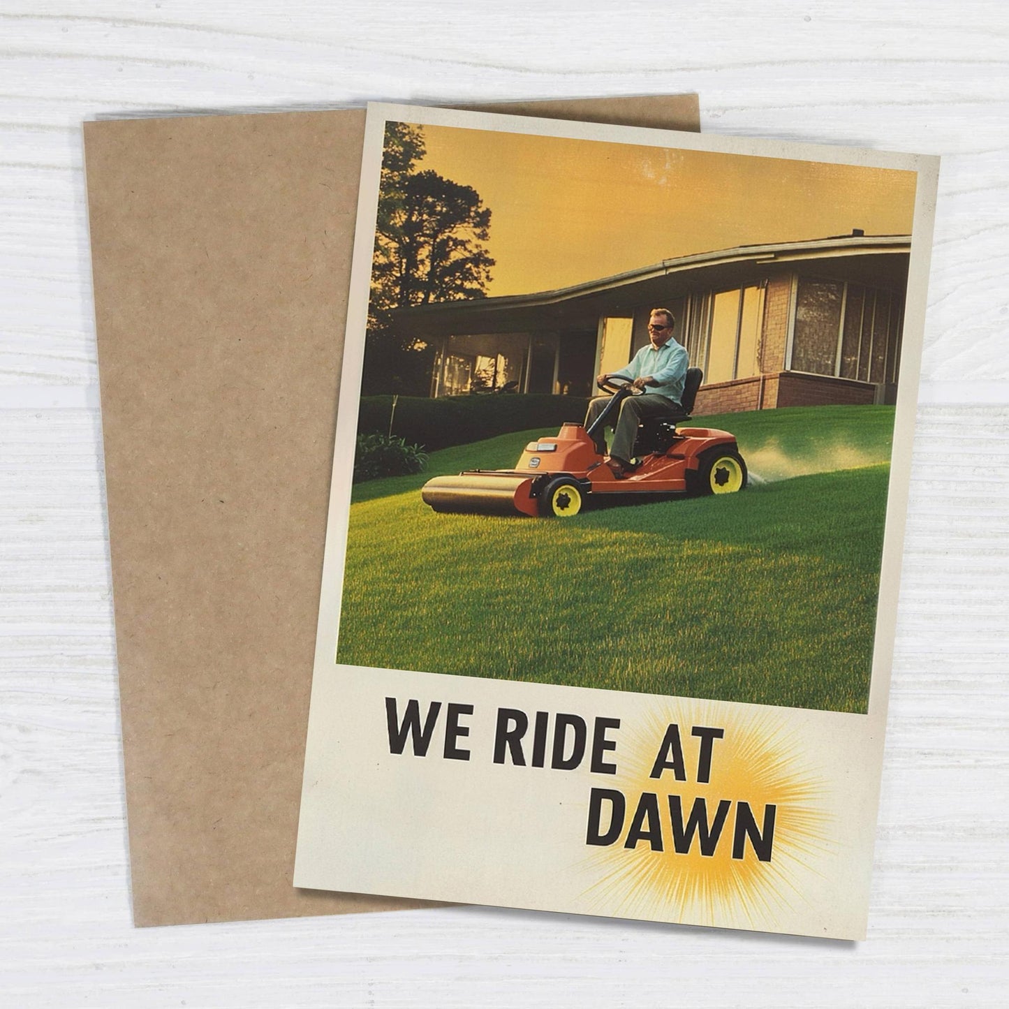 Greeting card with a man on a lawn mower and 'We Ride at Dawn' text, placed on a wooden surface.