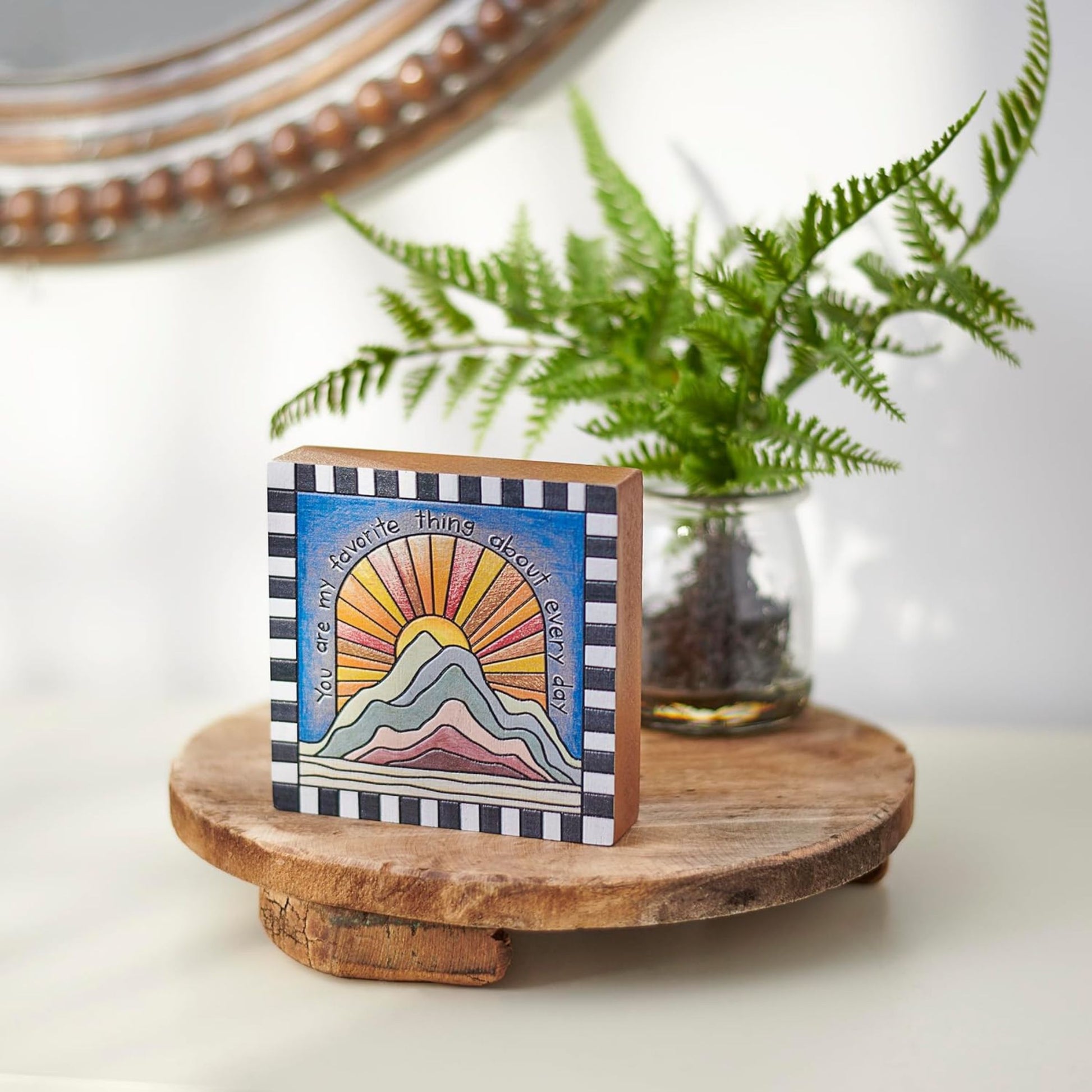 Small framed artwork with mountain and sun design on a wooden stand with a plant in the background.