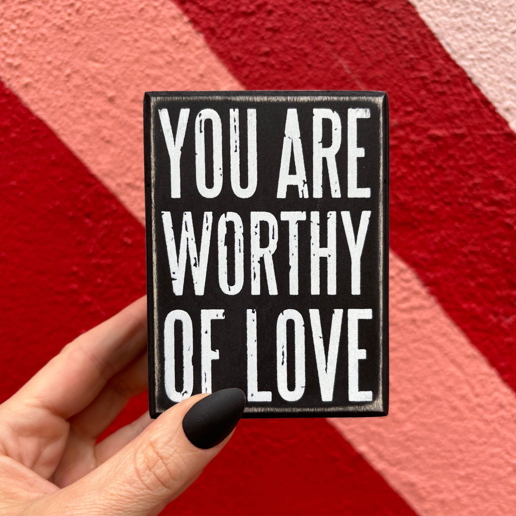 Black sign with 'YOU ARE WORTHY OF LOVE' text held against a red and pink striped background