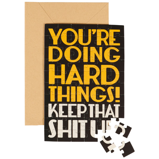Motivational card with yellow text 'You're doing hard things! Keep that shit up!' and a brown envelope on a white background.