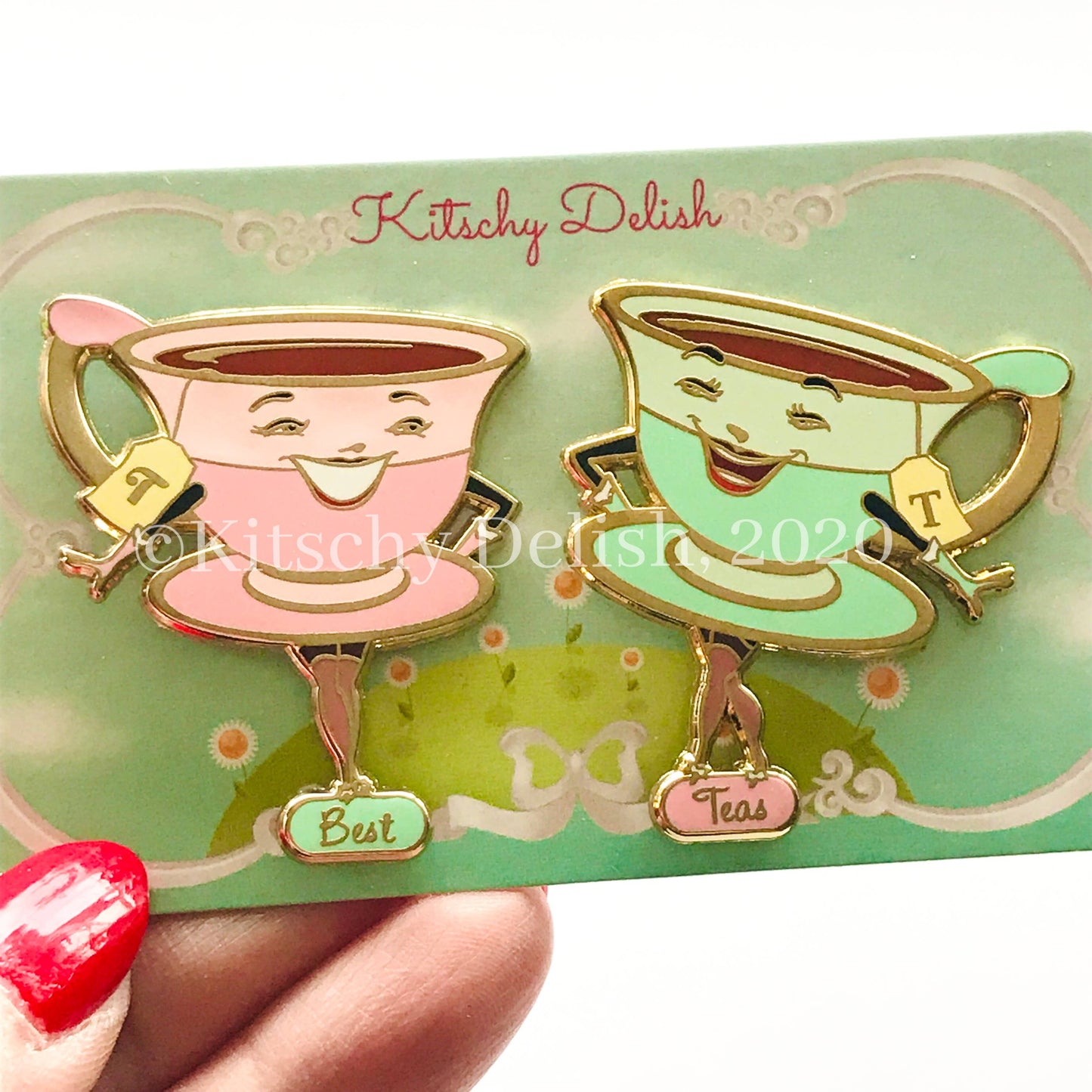 Two smiling teacup enamel pins on a green backing card, held by a hand with red nail polish. One cup is pink with gold trim and the word 'Best', and the other is mint green with gold trim and the word 'Teas'.