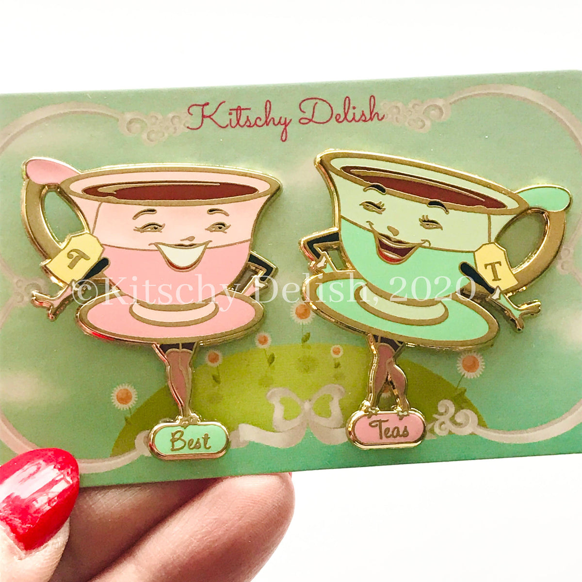 Two smiling teacup enamel pins on a green backing card, held by a hand with red nail polish. One cup is pink with gold trim and the word 'Best', and the other is mint green with gold trim and the word 'Teas'.