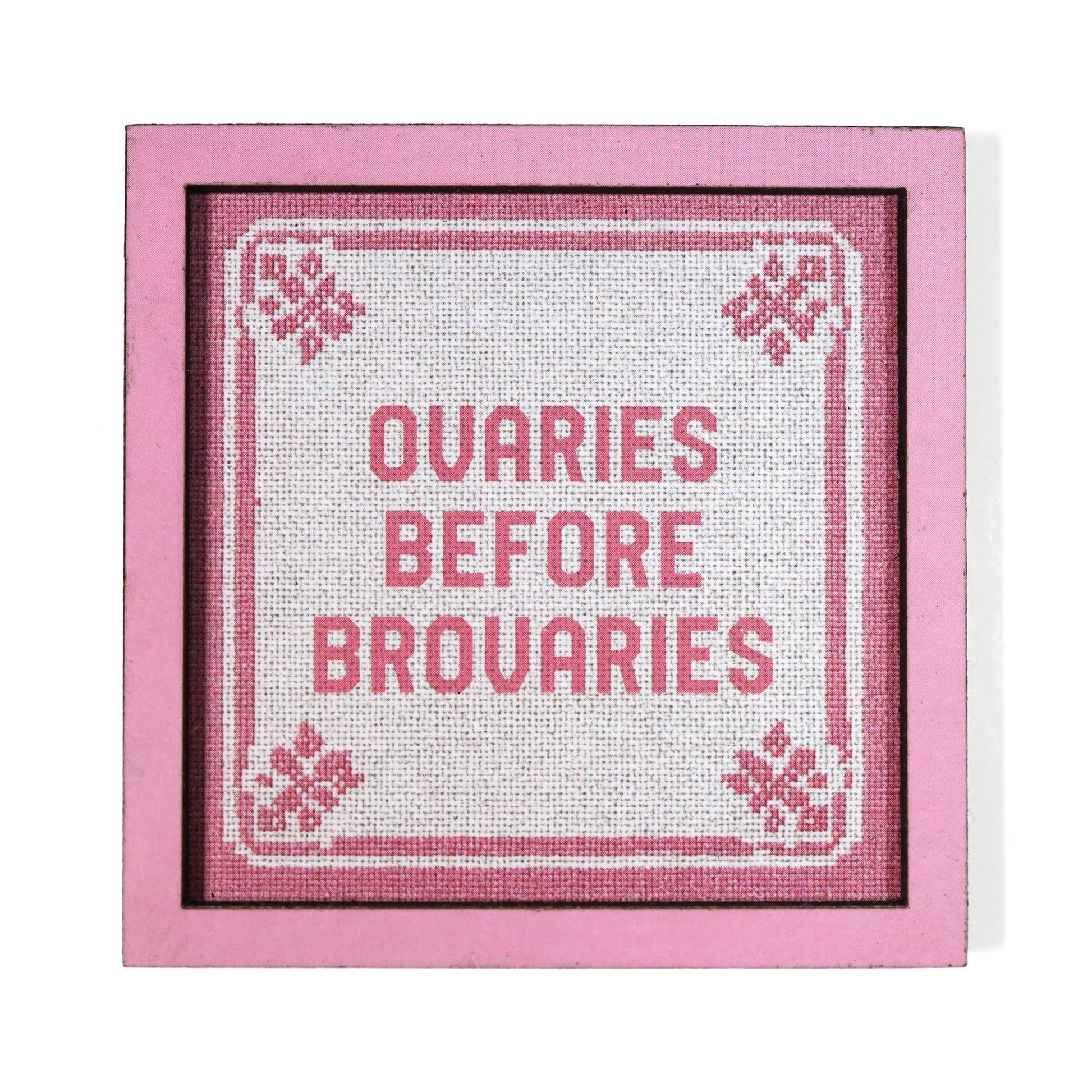 A square framed cross-stitch style magnet with pink border and text reading 'OVARIES BEFORE BROVARIES'