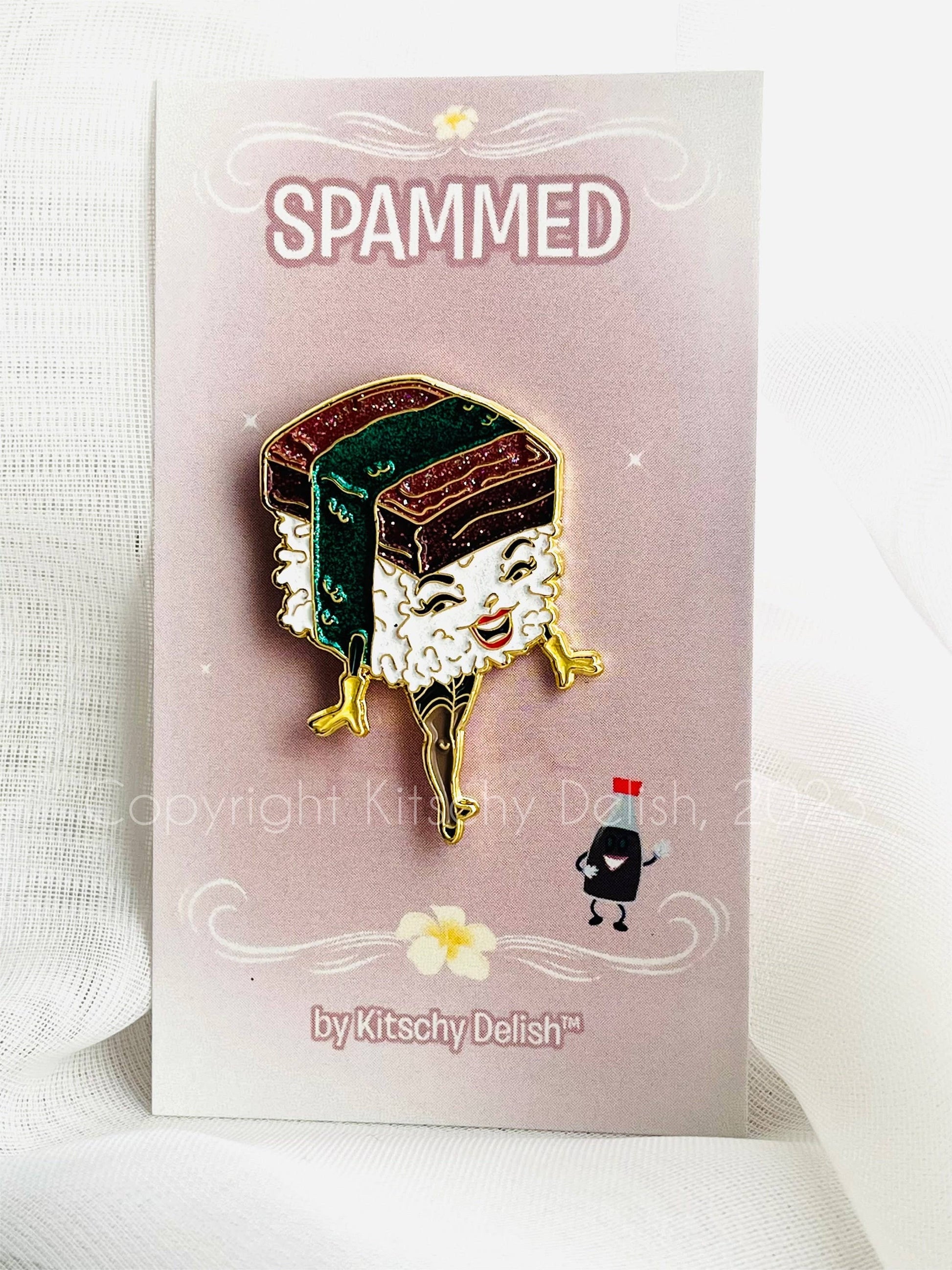 A cute spammed sushi enamel pin displayed on its pink backing card, which reads 'SPAMMED' and is branded 'by Kitschy Delish'. The pin features a smiling sushi character with white rice, green seaweed, and gold metal outlines.